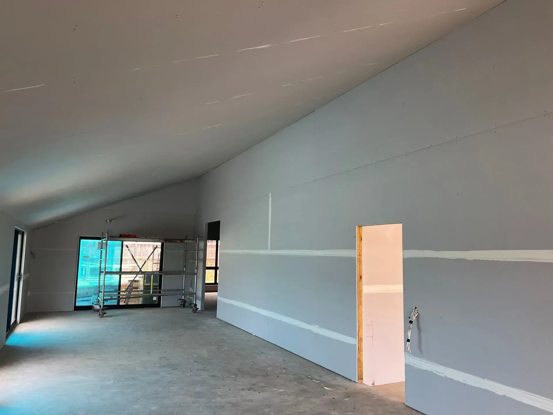 Empty unfinished room with sloped ceiling, gray walls, exposed flooring, and a doorway opening to scaffolding.