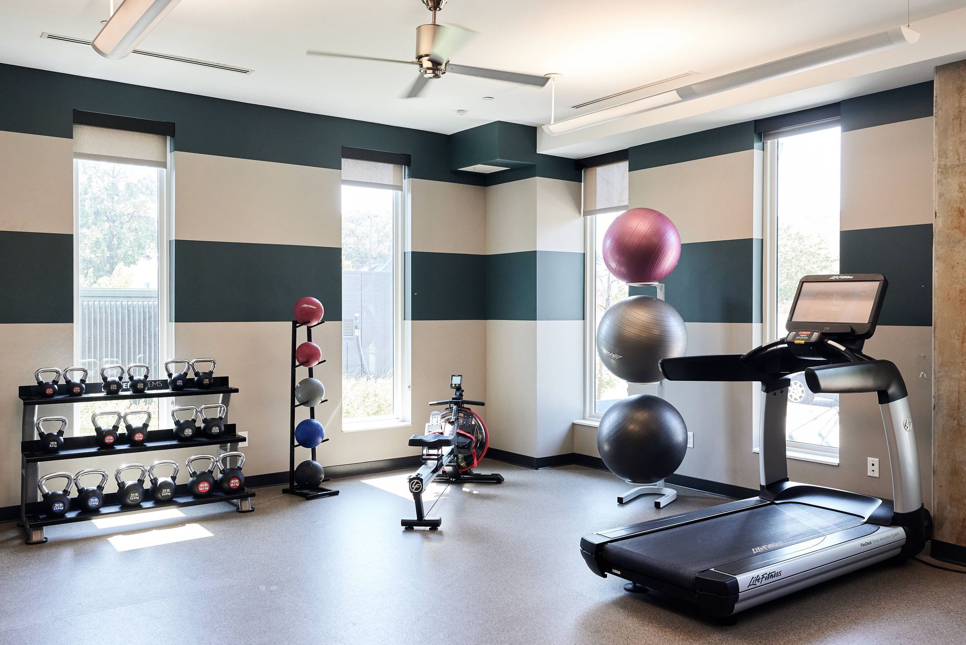 Gym with treadmill, kettlebells, exercise balls, stationary bike, and windows. Neutral walls, blue stripes.