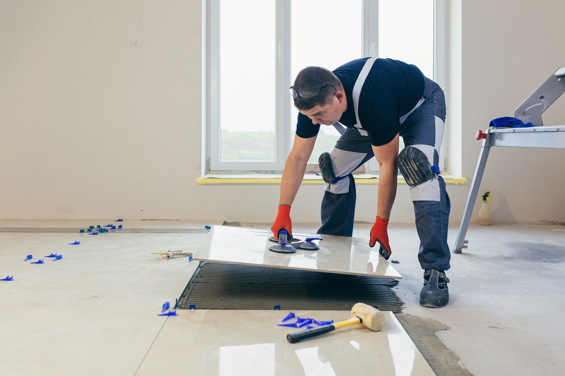 Man tiling a floor, kneeling with tools, wearing safety gear, in a room with a window.