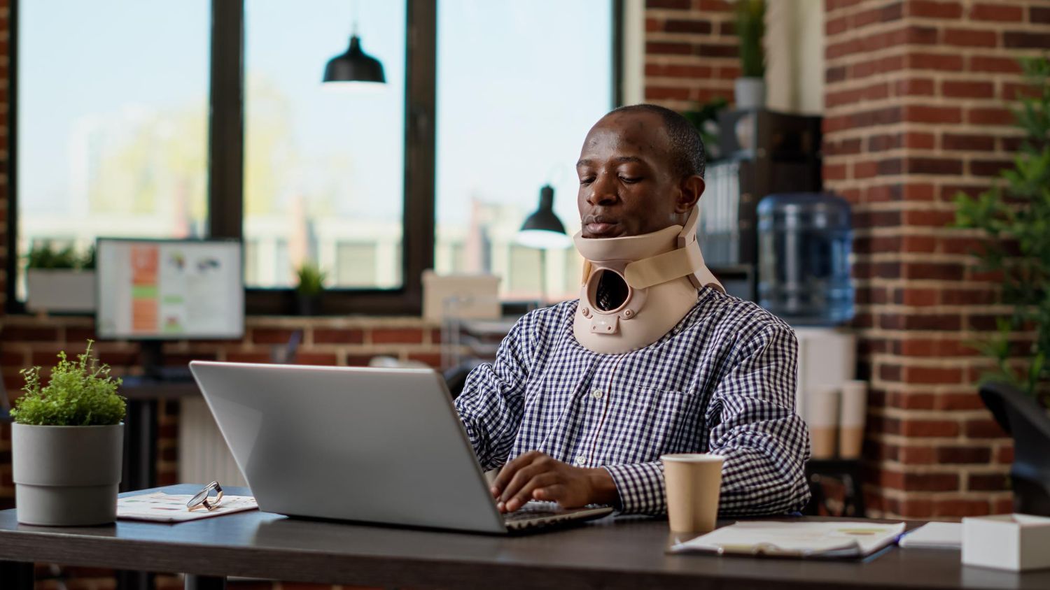 Person wearing a neck brace working on a laptop at a desk in a bright office