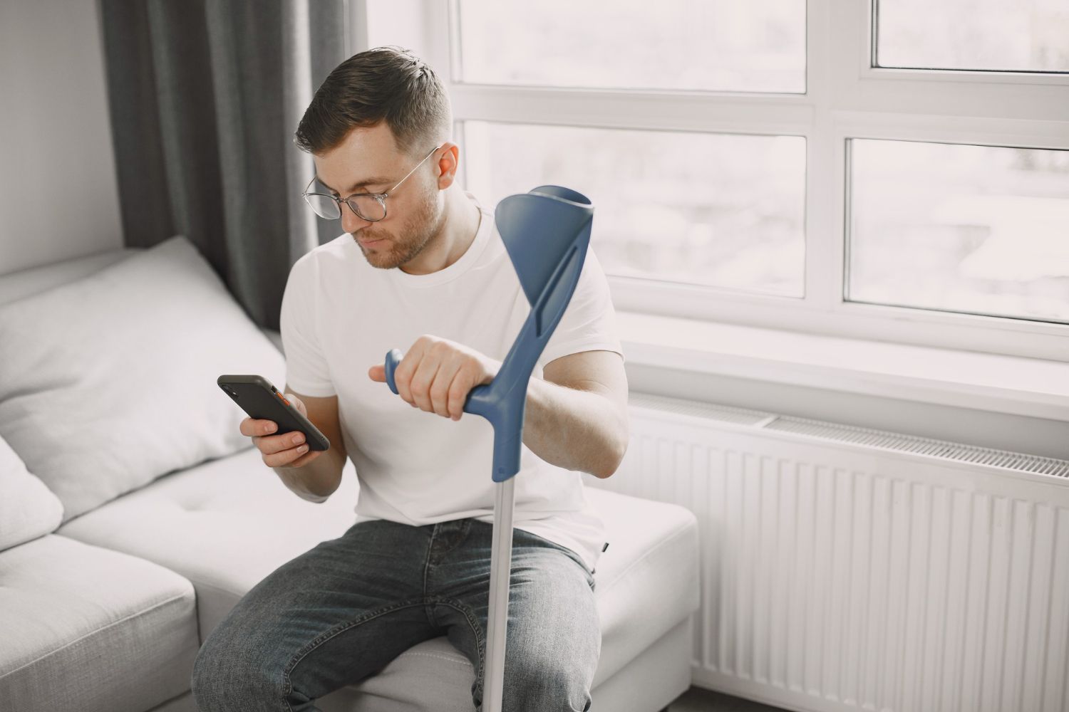 Man on bed using crutches while looking at a smartphone near a bright window