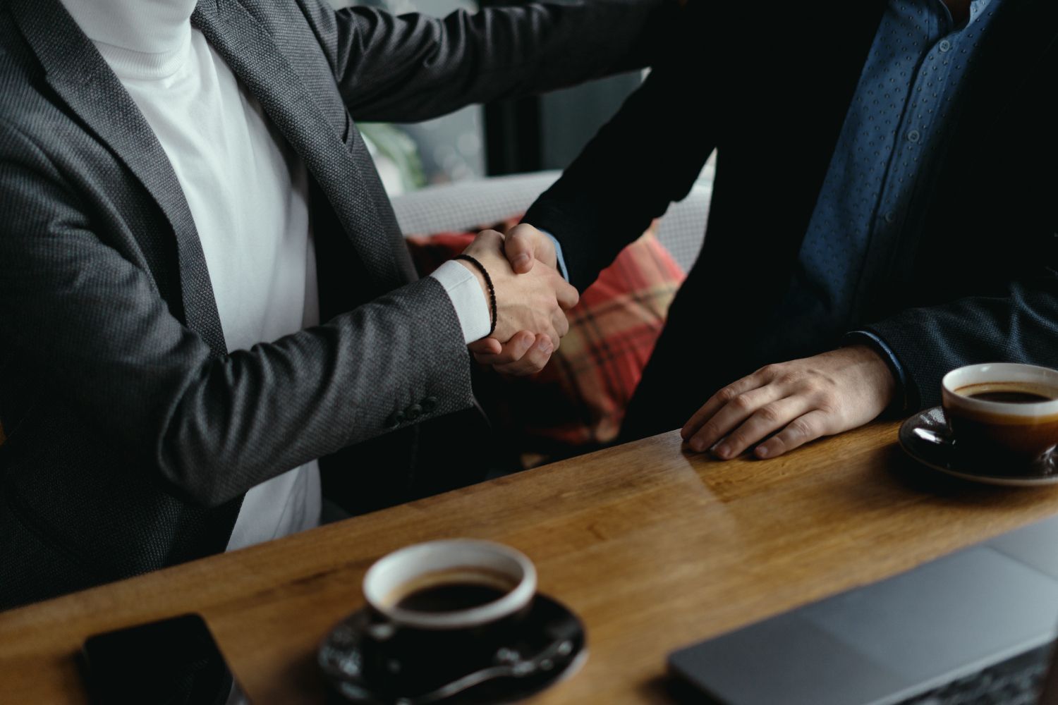 Two people shaking hands across a wooden table in a coffee shop, with cups of coffee nearby.