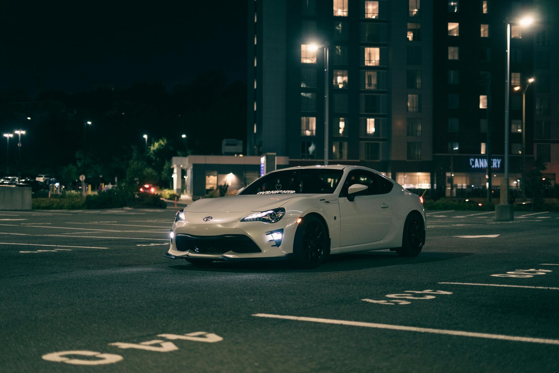 White sports car parked in a dimly lit city lot at night with apartment buildings in the background