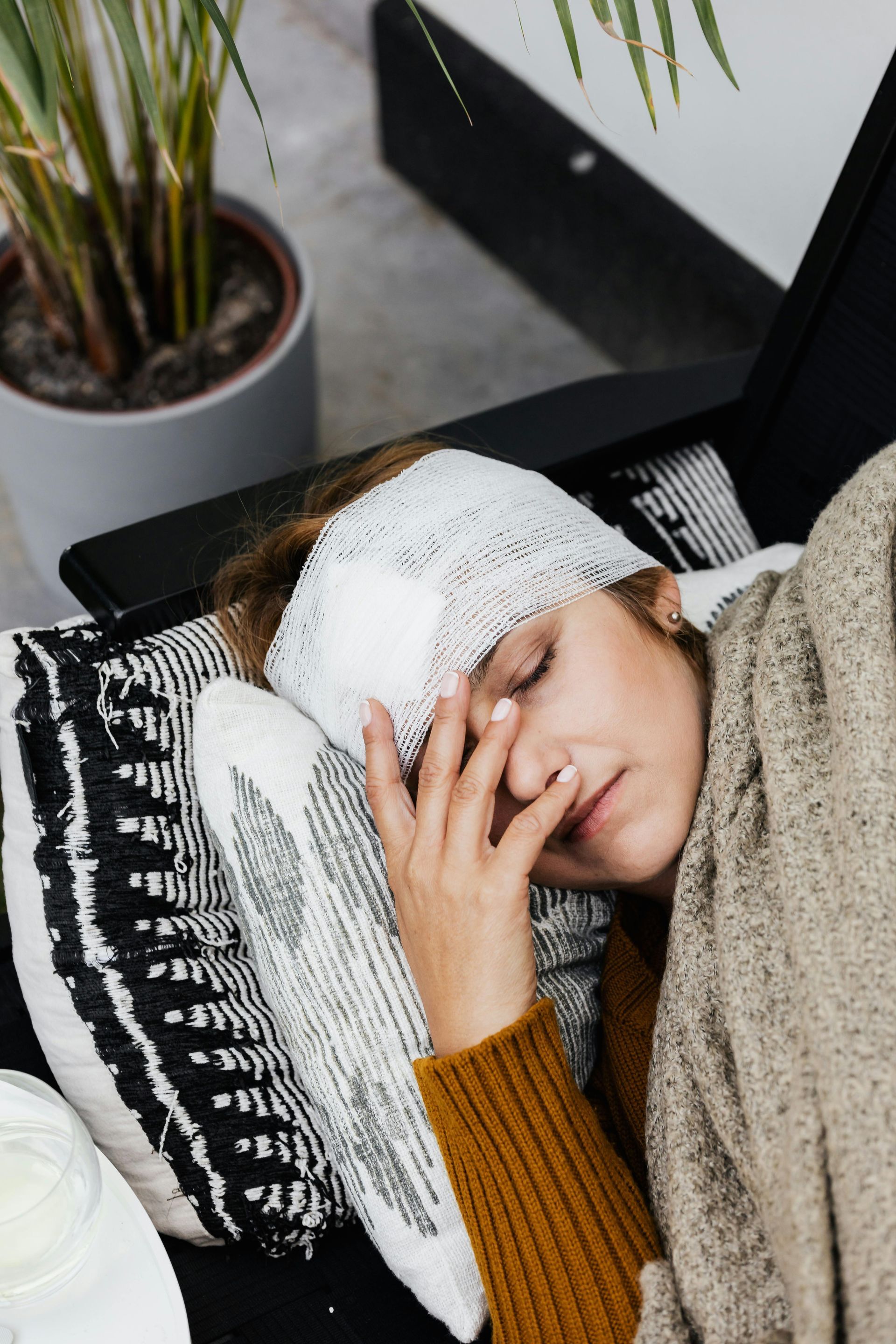 Person resting on a couch with a bandaged head, hand on face, under a beige blanket.