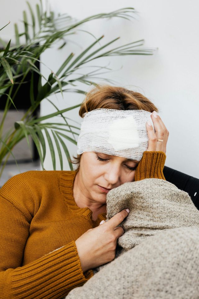 Person with head bandage resting on a beige blanket on a couch, wearing a mustard sweater.