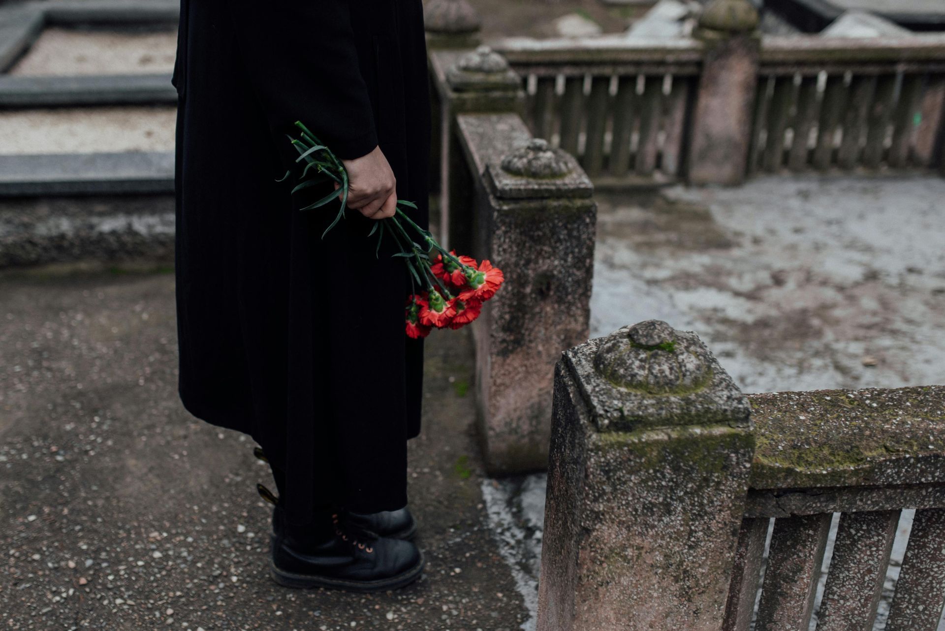 Person in a black coat holding red flowers beside a weathered stone bench outdoors