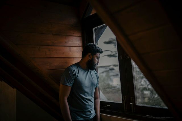 Man standing by a window in a dim wooden attic, looking outside.