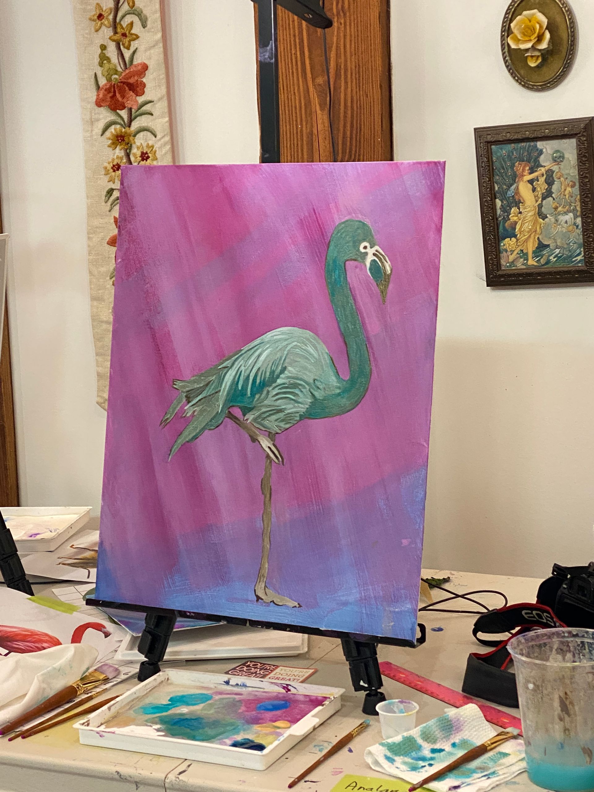 Painting of a teal flamingo on a purple canvas in an art studio, surrounded by paint supplies and sketches