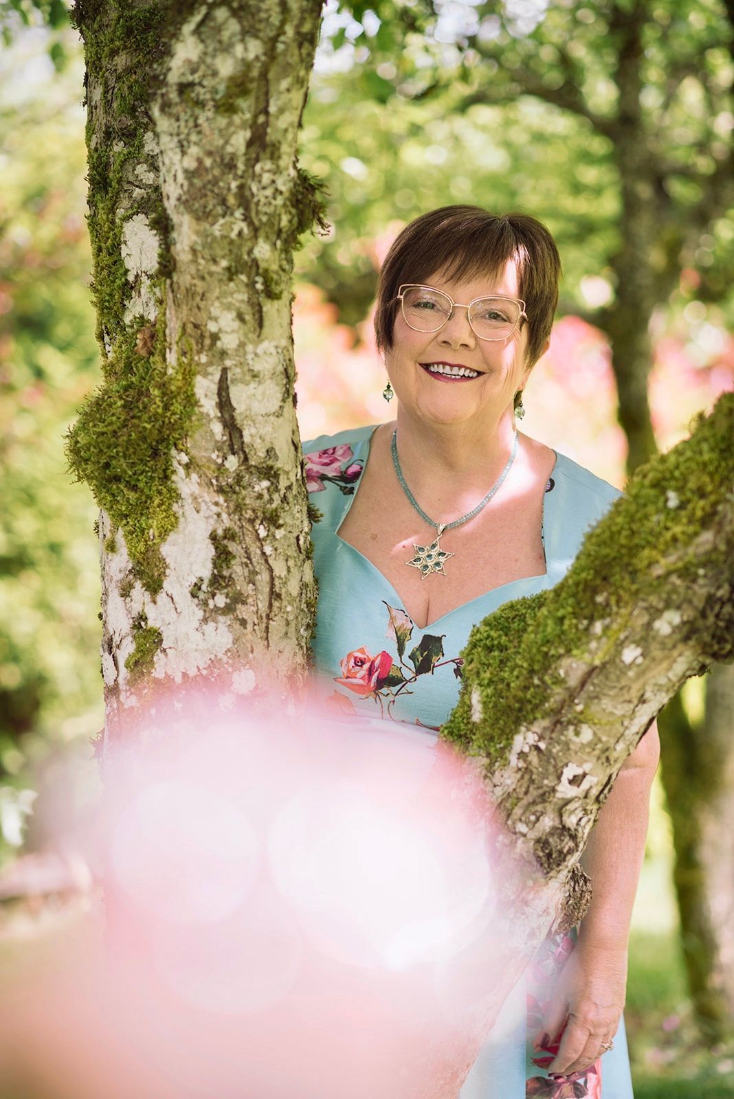 Woman smiling between mossy tree trunks in a sunlit garden, with soft pink blur in the foreground