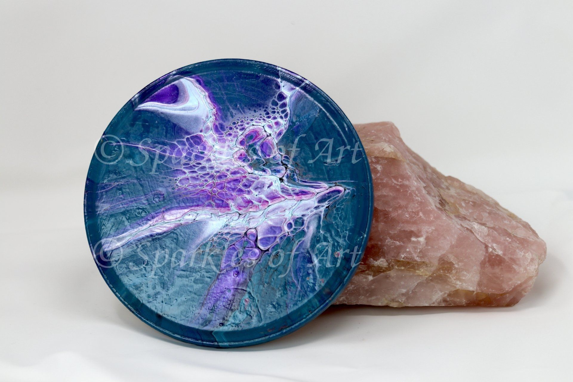 Blue circular crystal-like disc with purple-white abstract design, resting against a pink stone on a white background