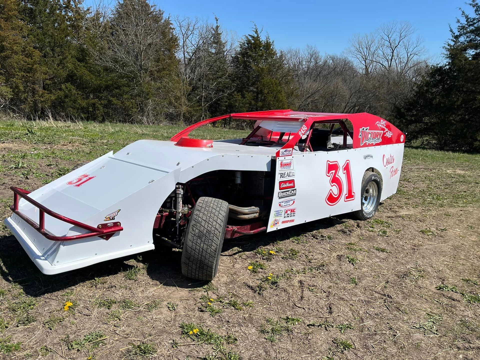 Red and white race car with number 31 parked on grass, front hood open outdoors