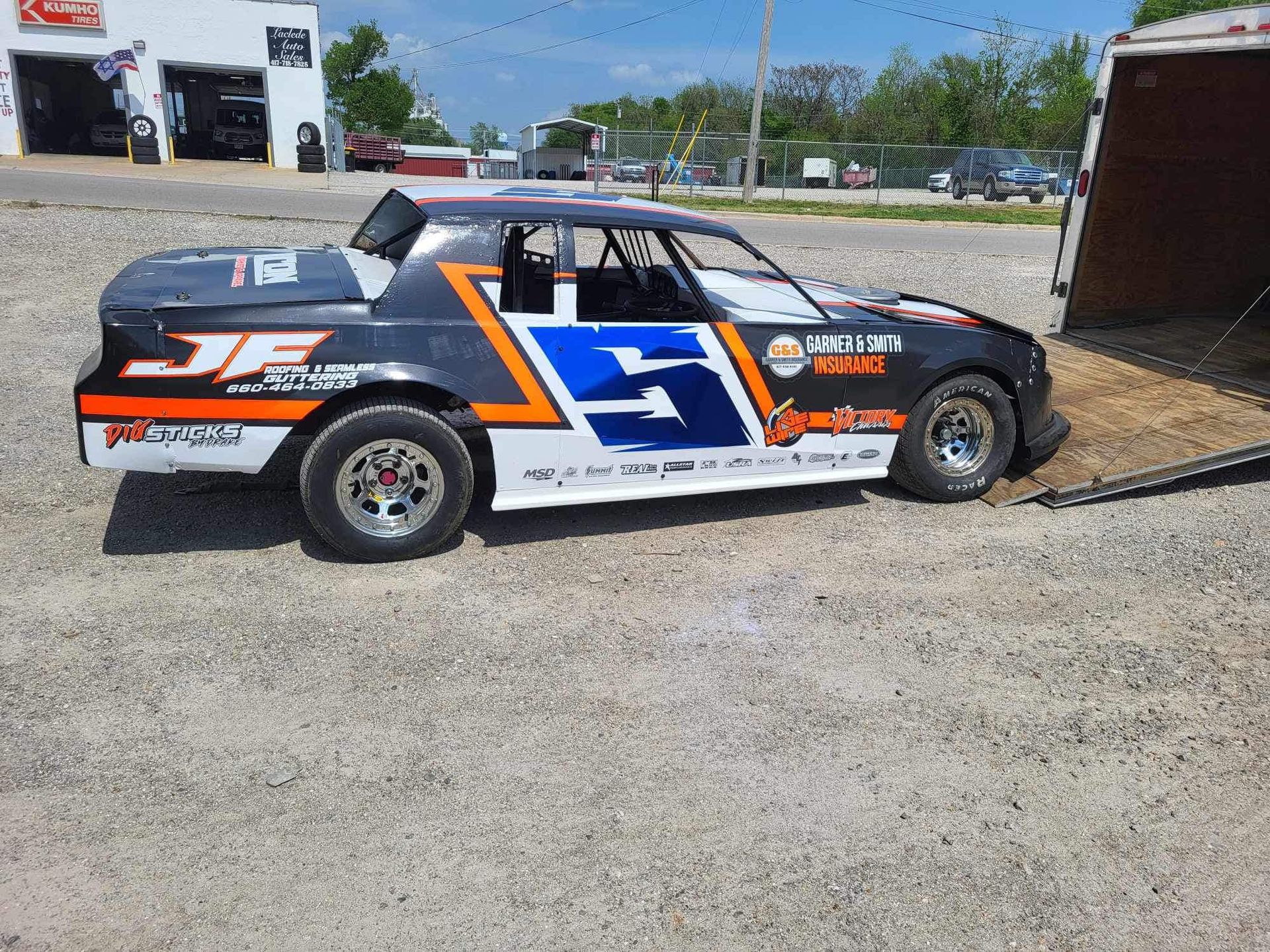 Race car with number 5 in blue and orange, parked on gravel near a trailer under a bright sky