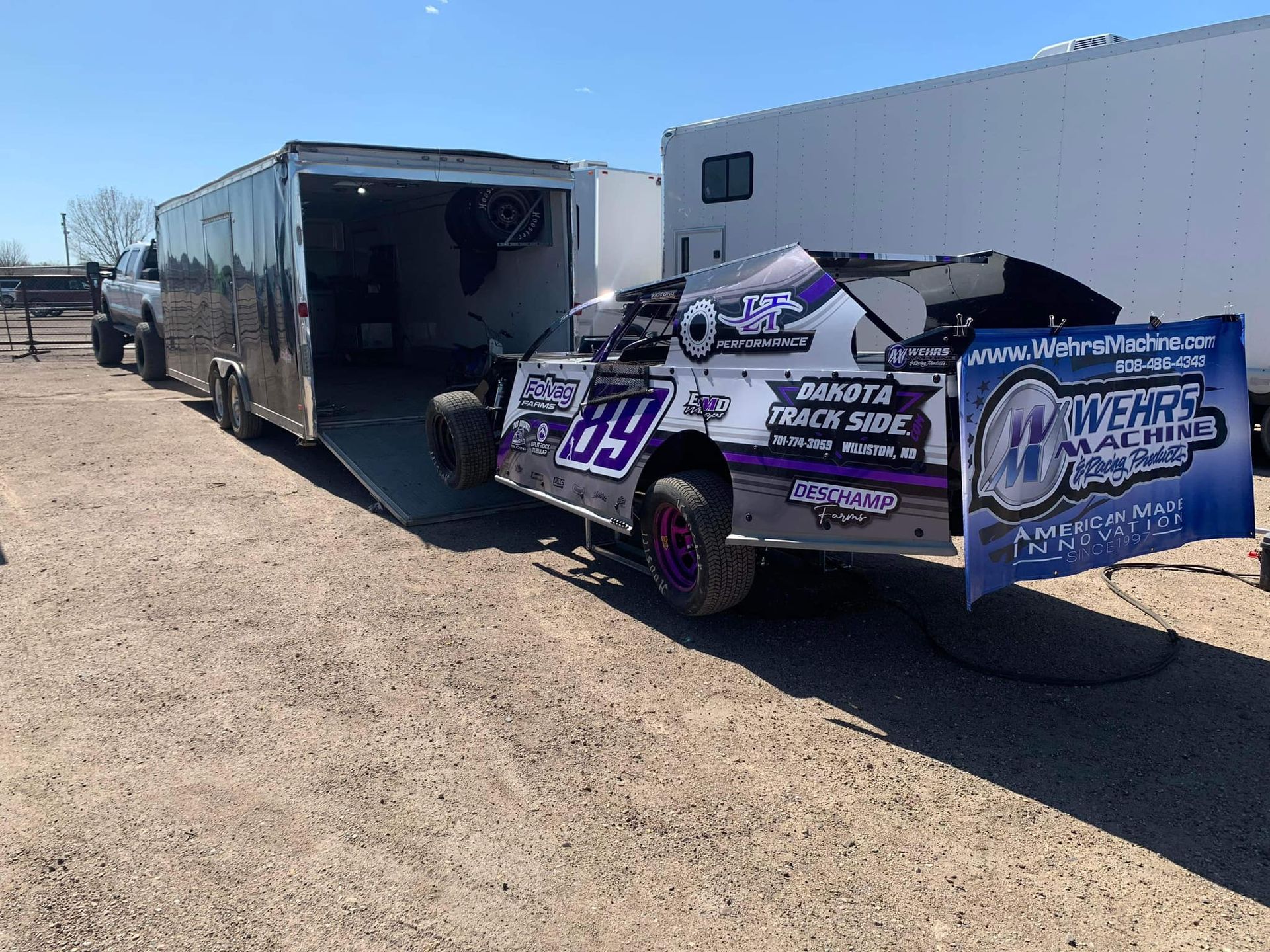 Race car parked beside an open trailer in a dirt lot, with a white trailer and blue sponsor graphics.