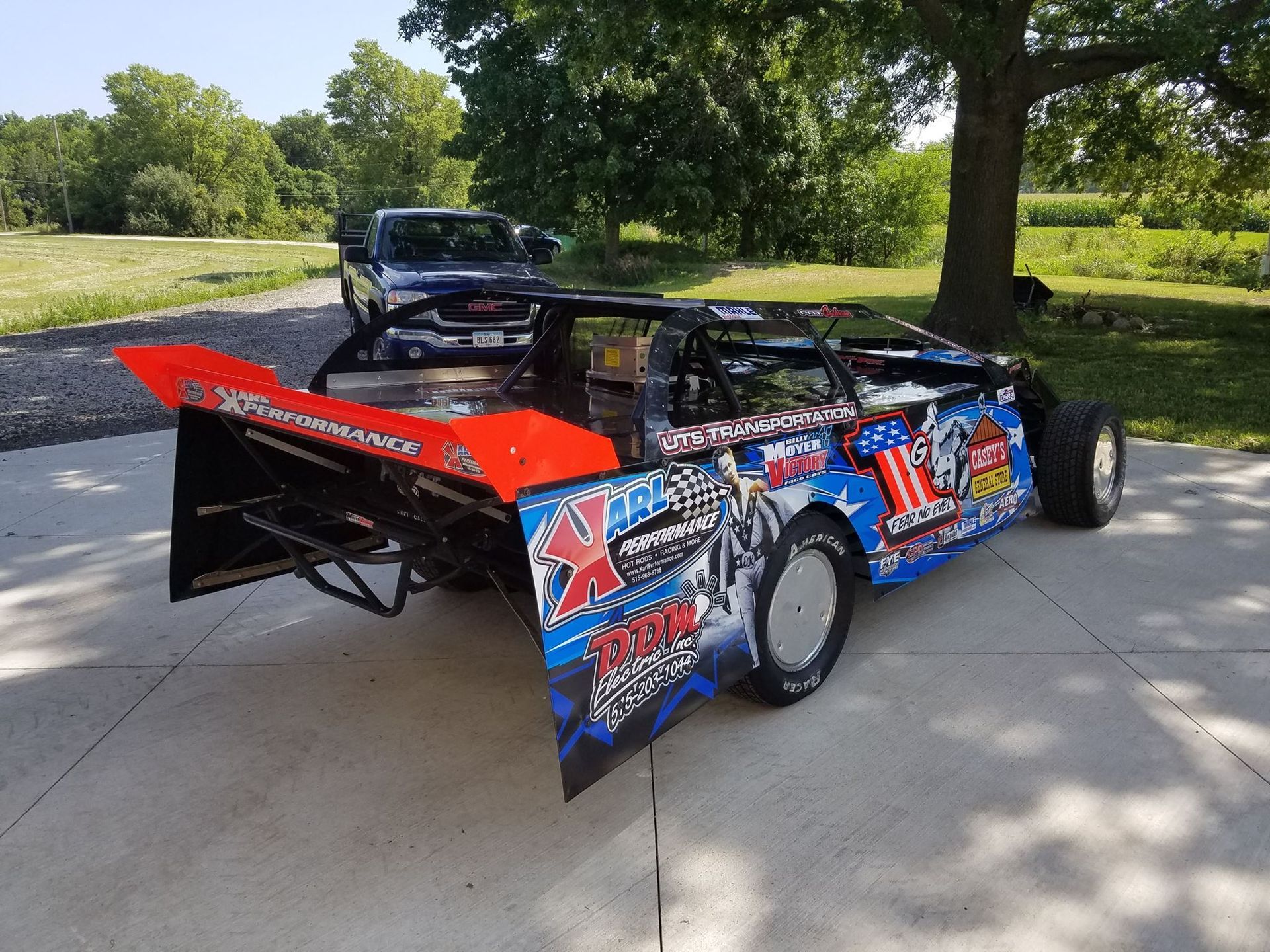 Blue and red dirt race car with sponsor decals parked on a driveway in a sunny wooded area