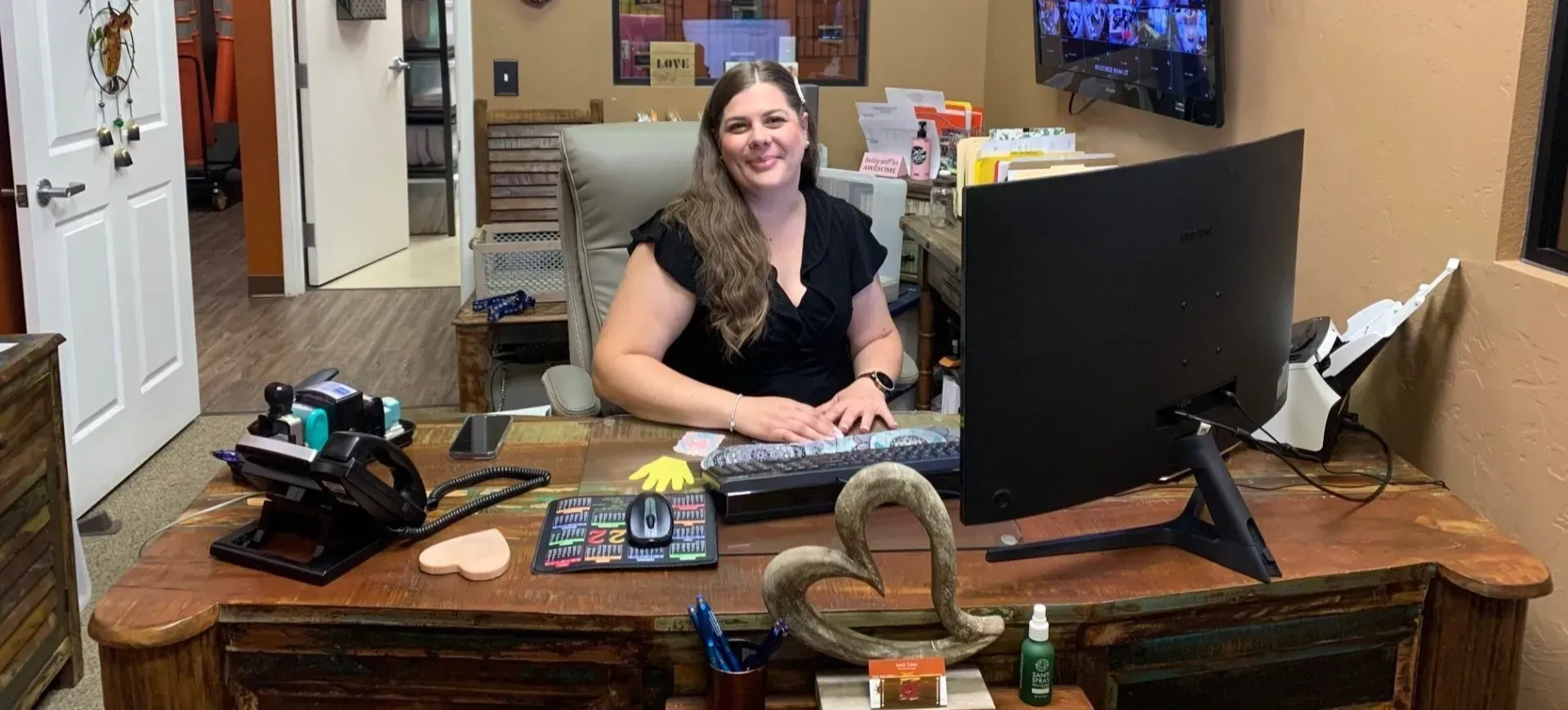 Woman seated at a desk, smiling. She is in an office with a computer and phone, working.