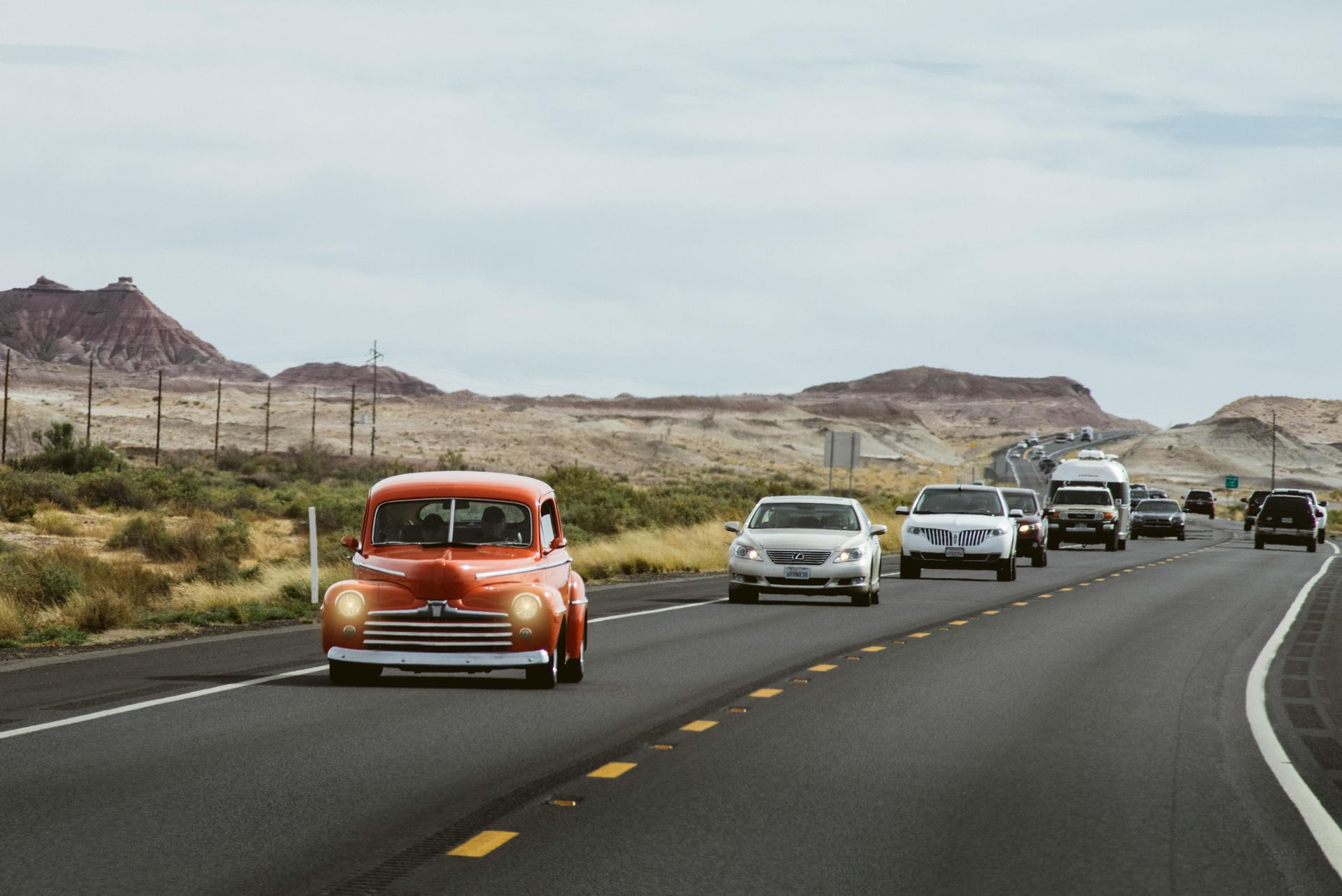 Orange vintage car on a highway with other vehicles, driving toward a desert landscape under a cloudy sky.