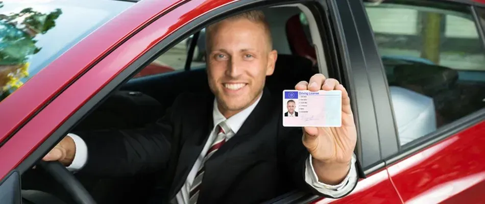 Man in a suit holding up a driver's license inside a red car. He is smiling.