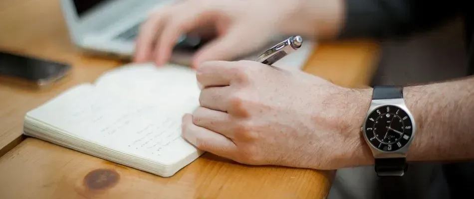 Person writing in a notebook, with a watch on their wrist, and laptop.
