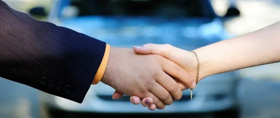Two hands shaking in front of a car, suggesting a deal. One hand is in a suit, the other has a bracelet.