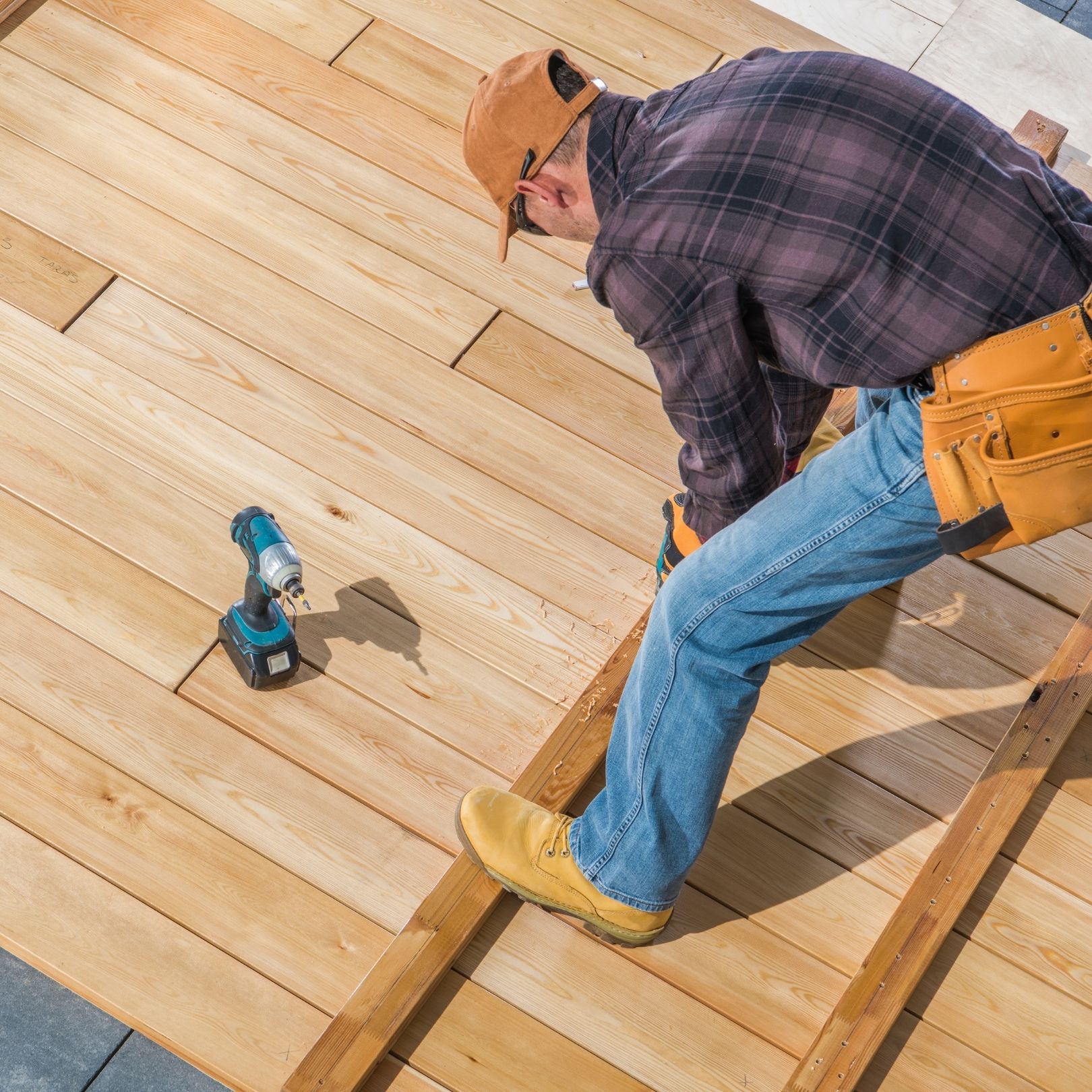 Worker kneeling on wooden deck boards, using a power nailer to install flooring.