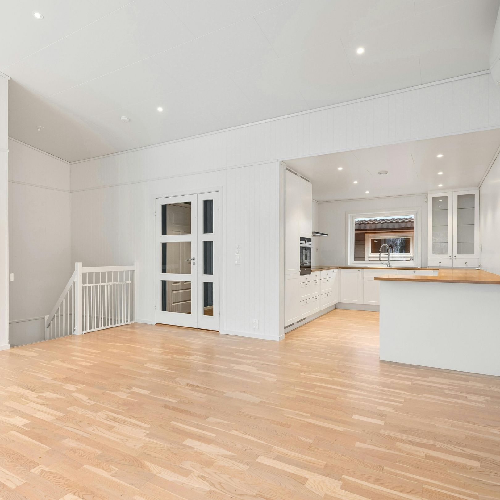 Bright, empty white living room with light wood floors and an open kitchen pass-through.