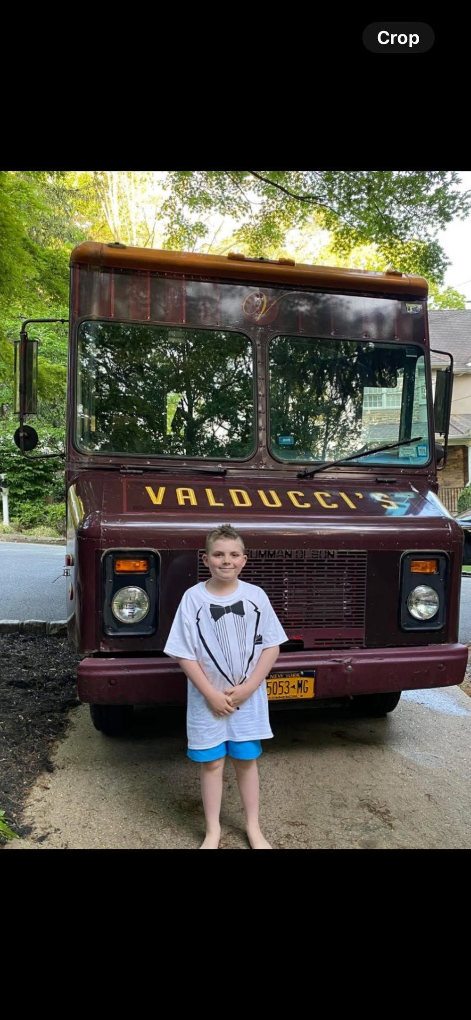 Child standing in front of a maroon truck labeled “WALDORF” in a wooded area
