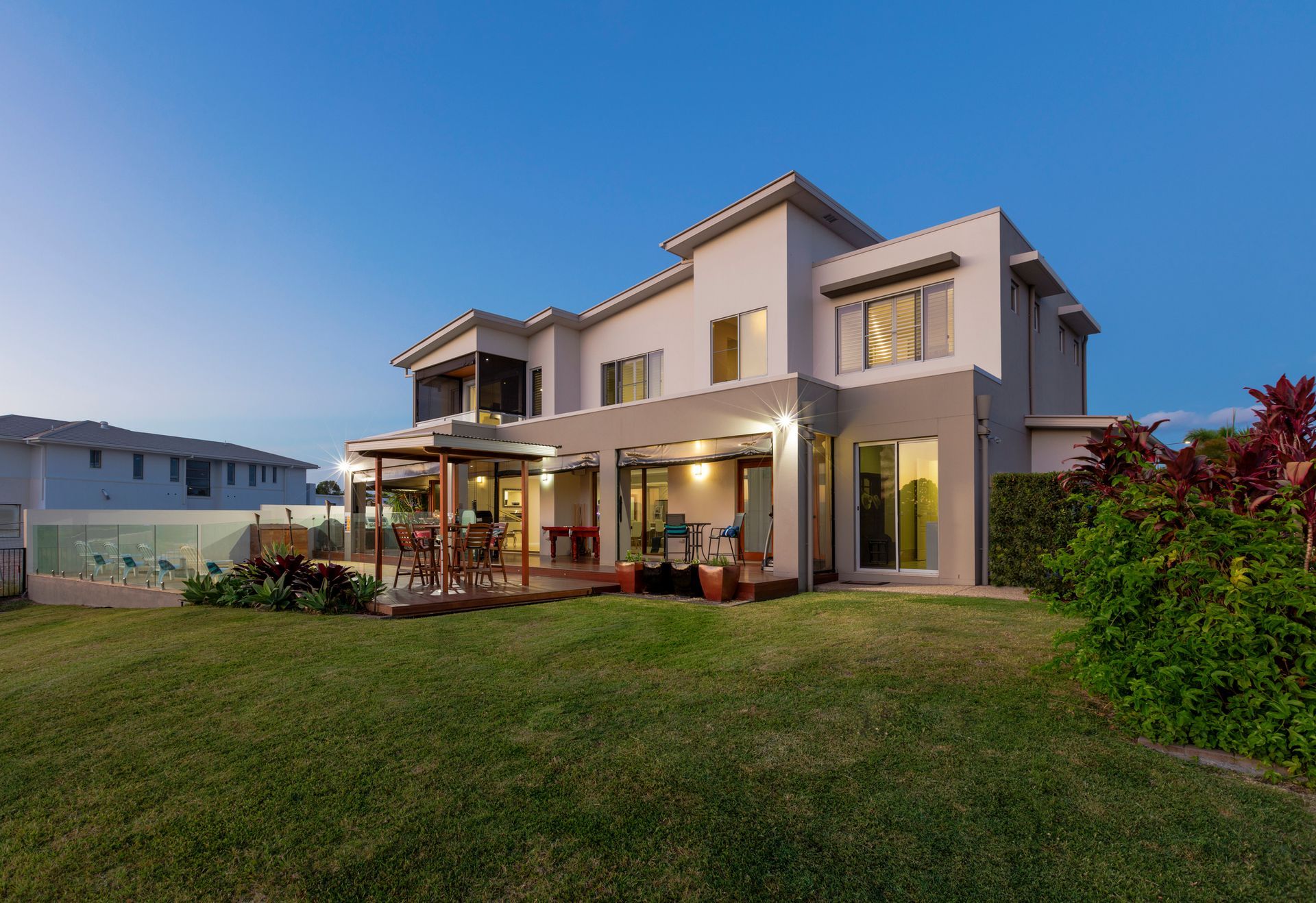 A modern two-story house at dusk featuring large windows, an outdoor patio with seating, a covered dining area A modern two-story house at dusk featuring large windows, an outdoor patio with seating, a covered dining area