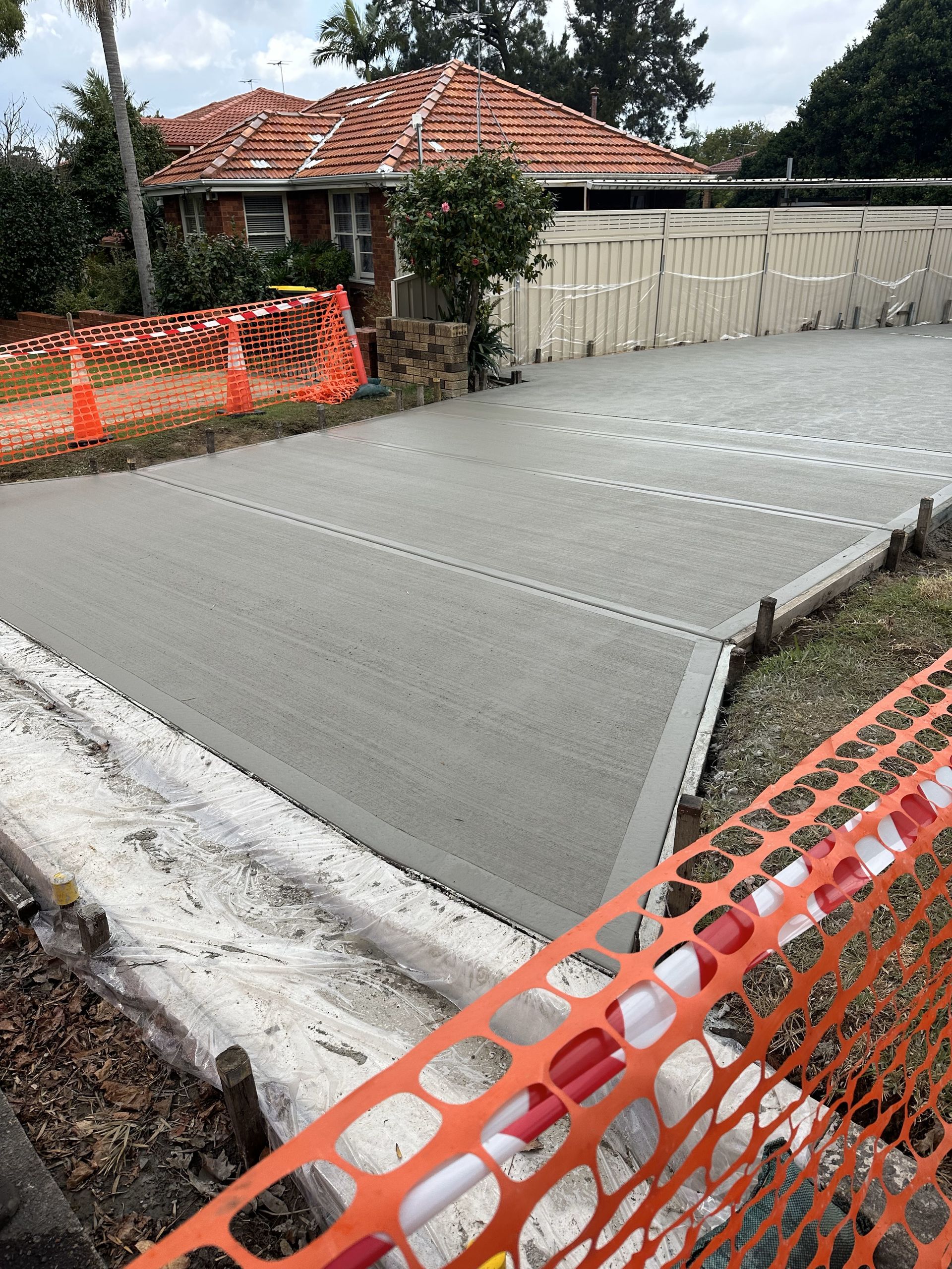 Freshly poured concrete driveway bordered by orange construction fencing. A house is in the background.