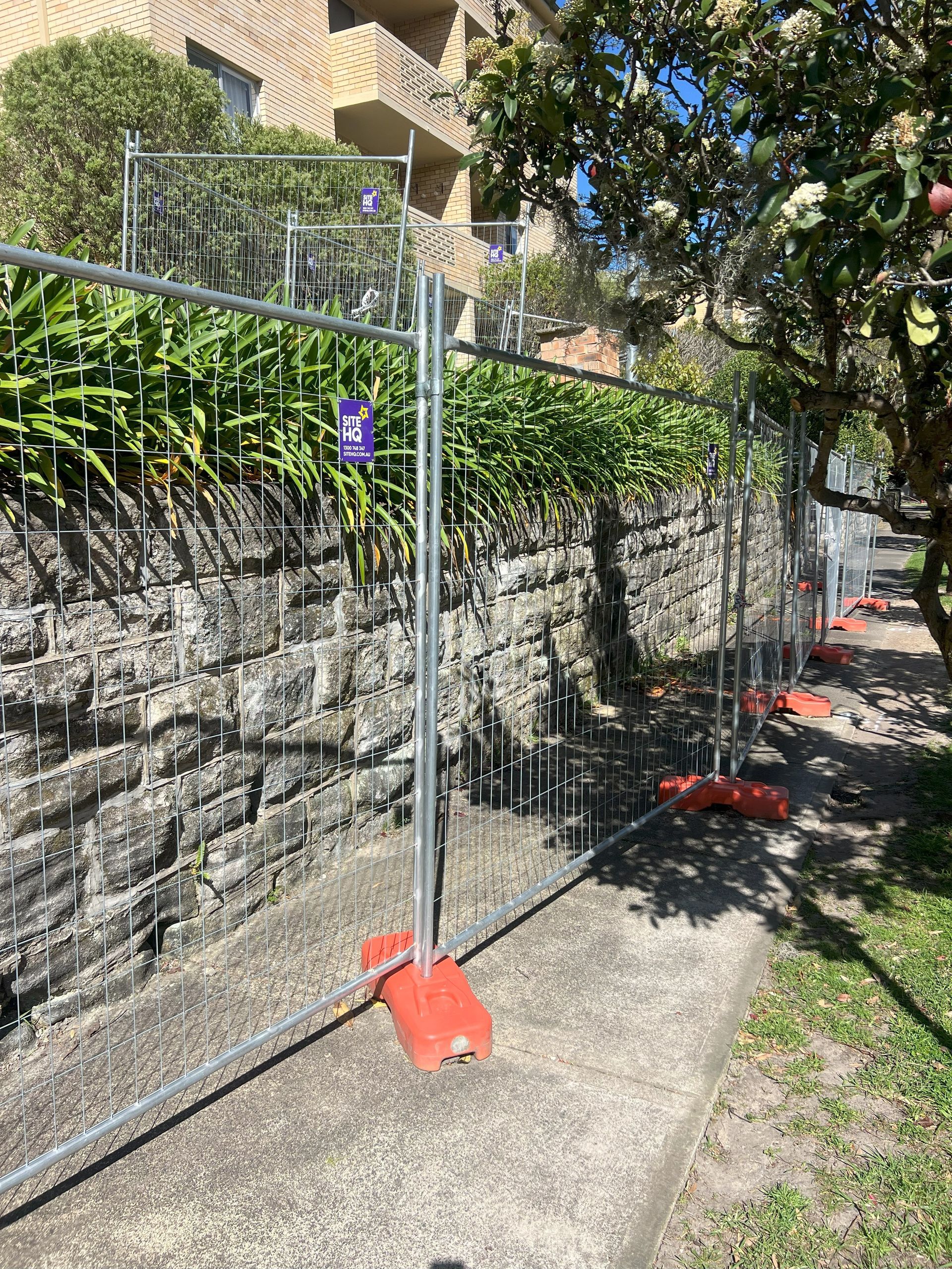 Temporary fence along a stone wall and sidewalk. Orange bases support the metal barriers.
