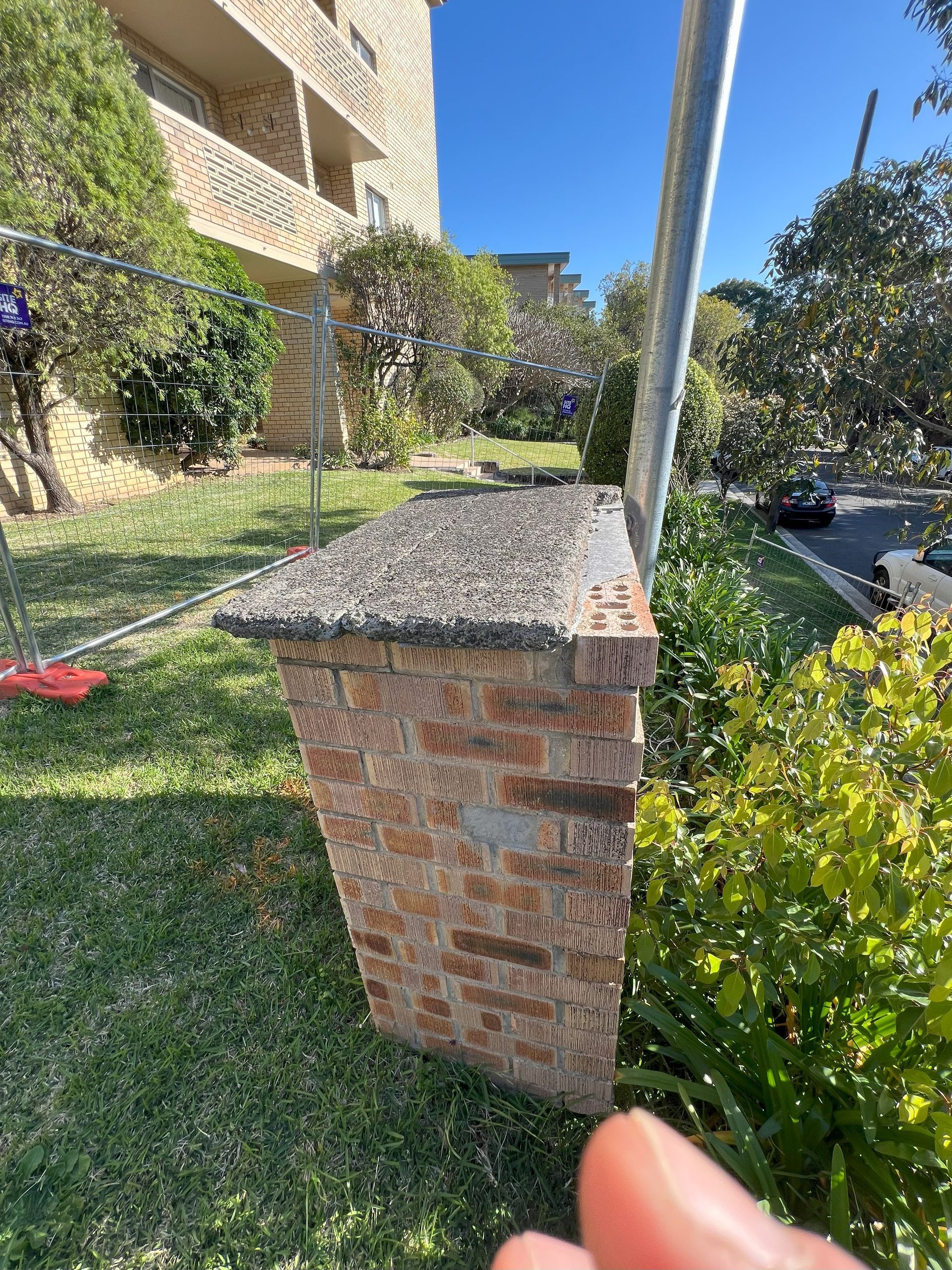 Brick pillar topped with rough concrete, beside a green lawn and building, pole at the edge, finger in the foreground.