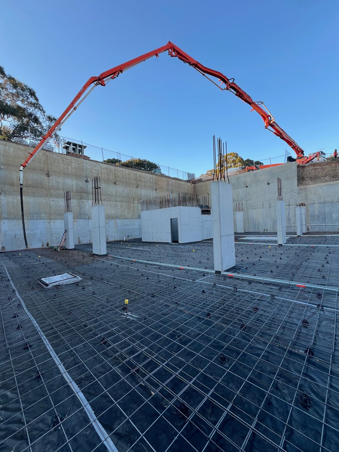 Construction site: Concrete pump extending over a rebar-covered foundation, concrete pillars, retaining wall in the background, blue sky.