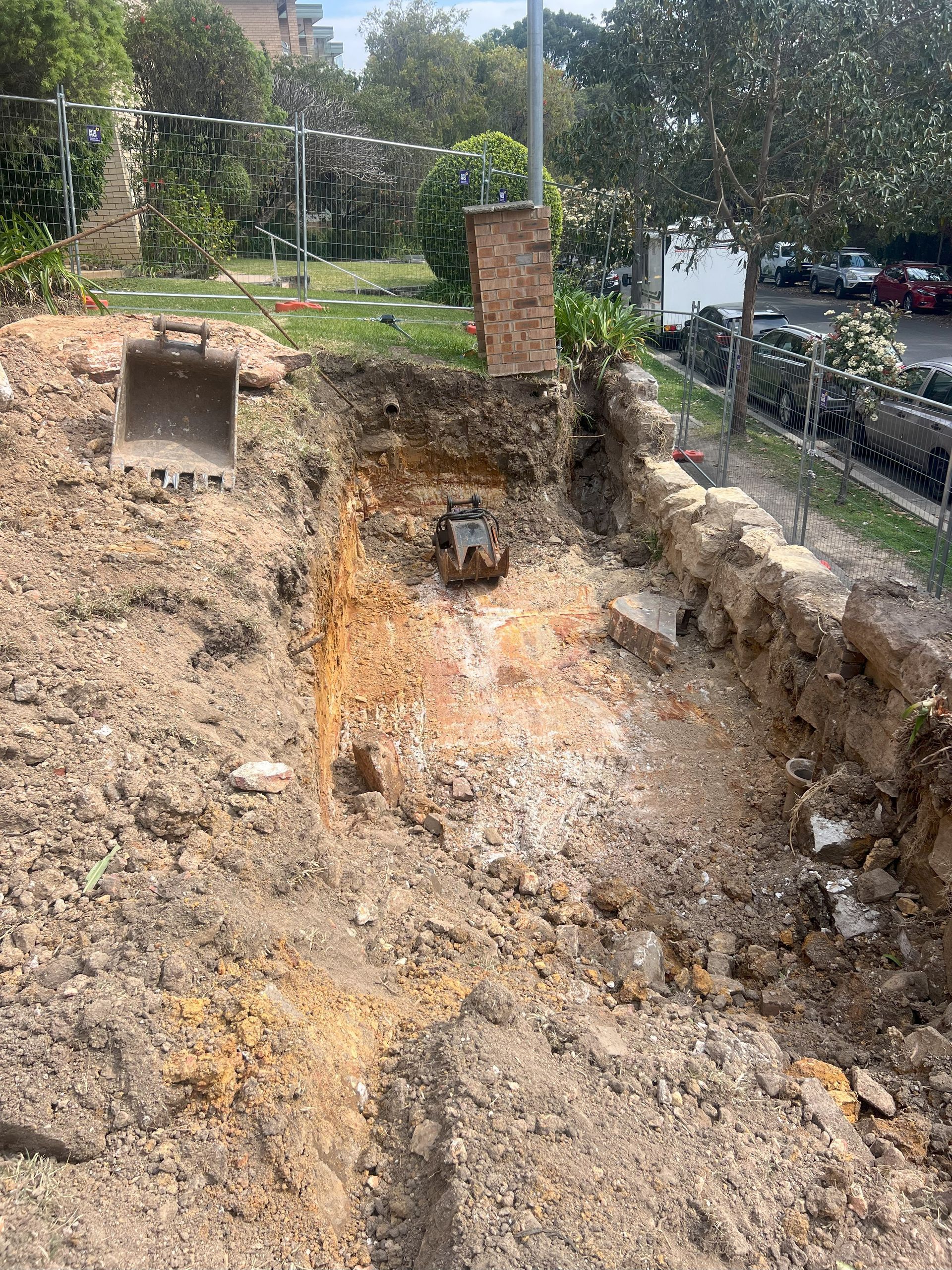 Construction site excavation. An excavator sits in the dirt pit. Soil and brick remain.