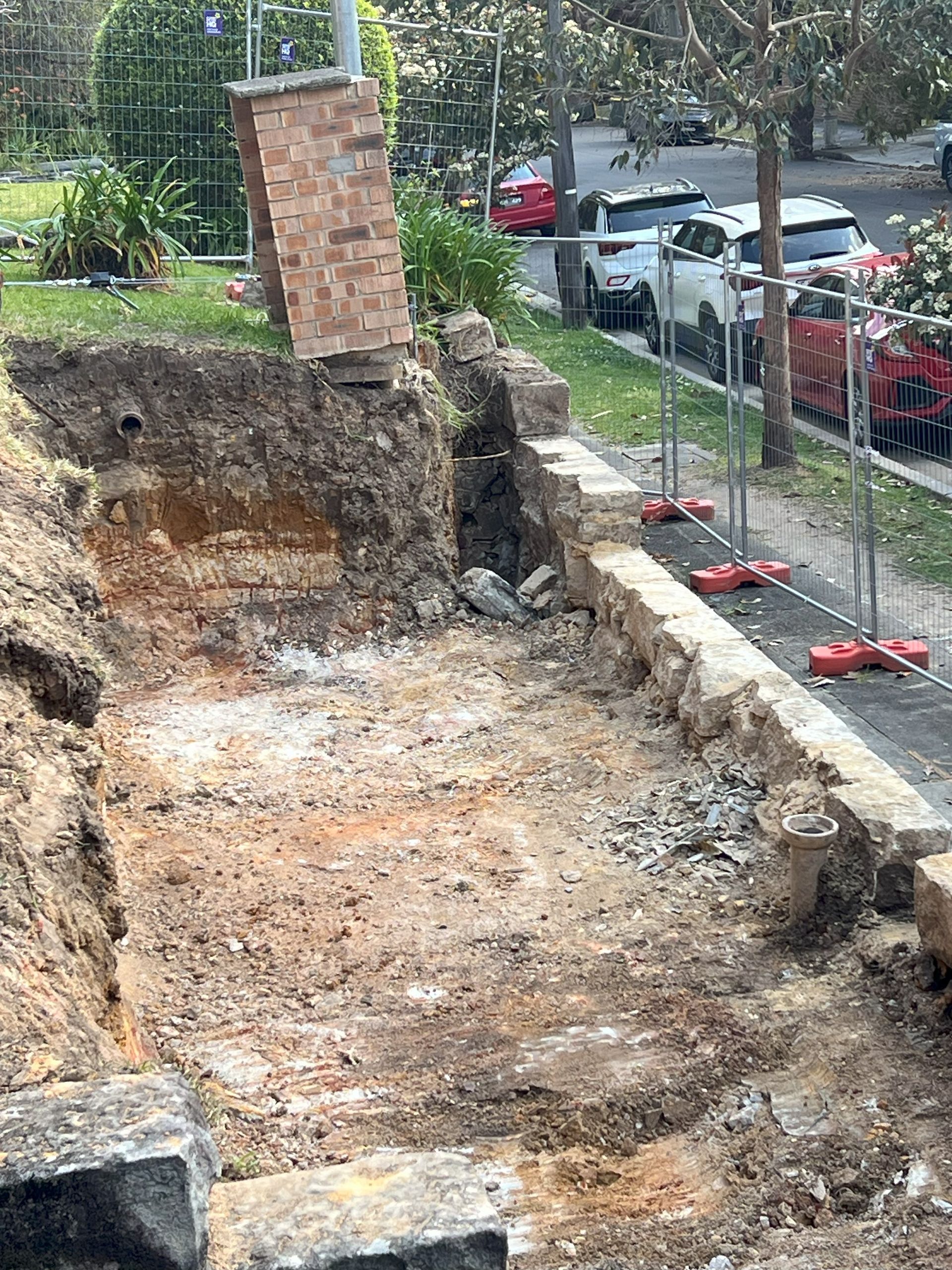 Excavation beside a street with a brick structure, retaining wall, dirt, safety fencing, and parked cars.