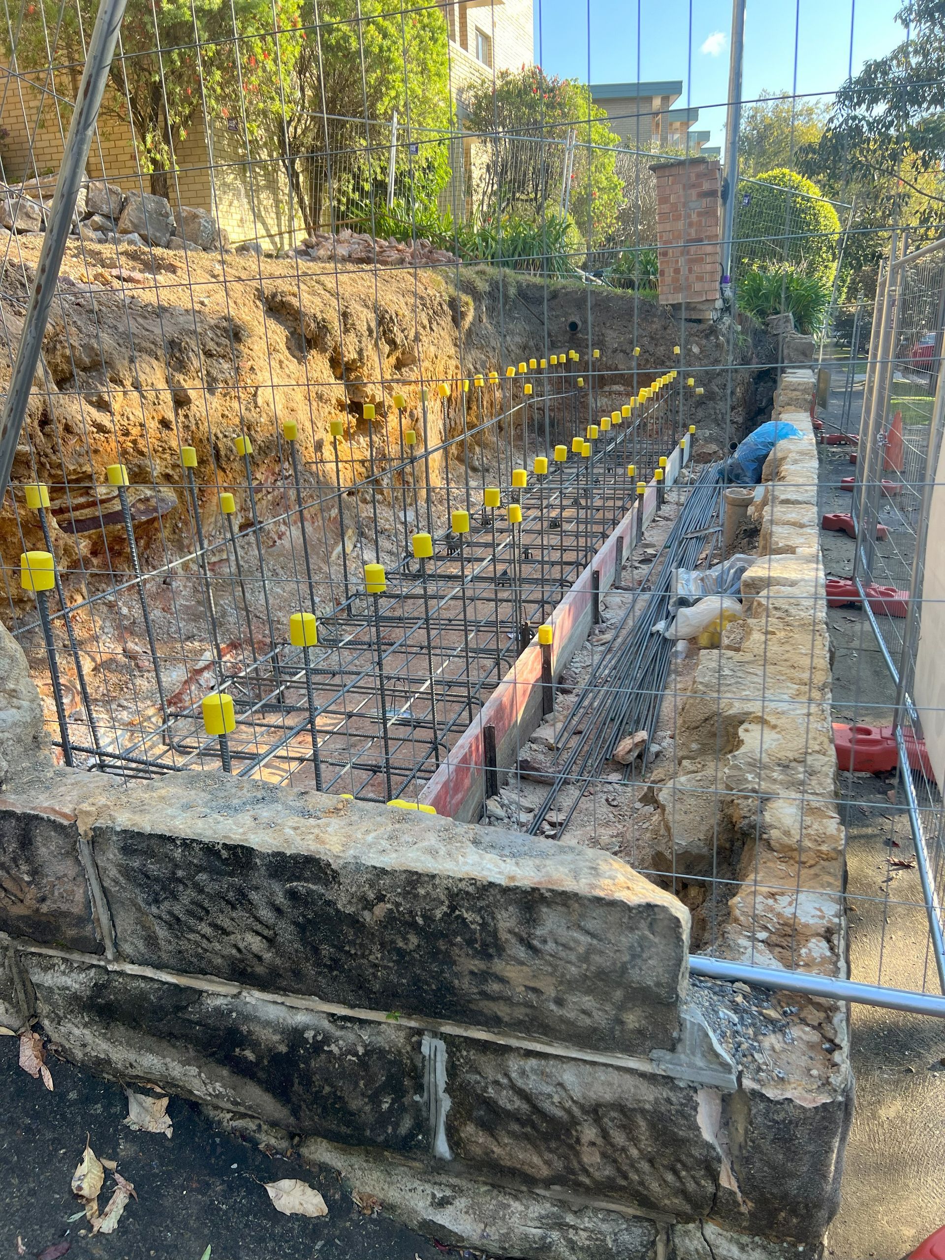 Construction site with exposed rebar, concrete forms, and a worker near a stone wall.