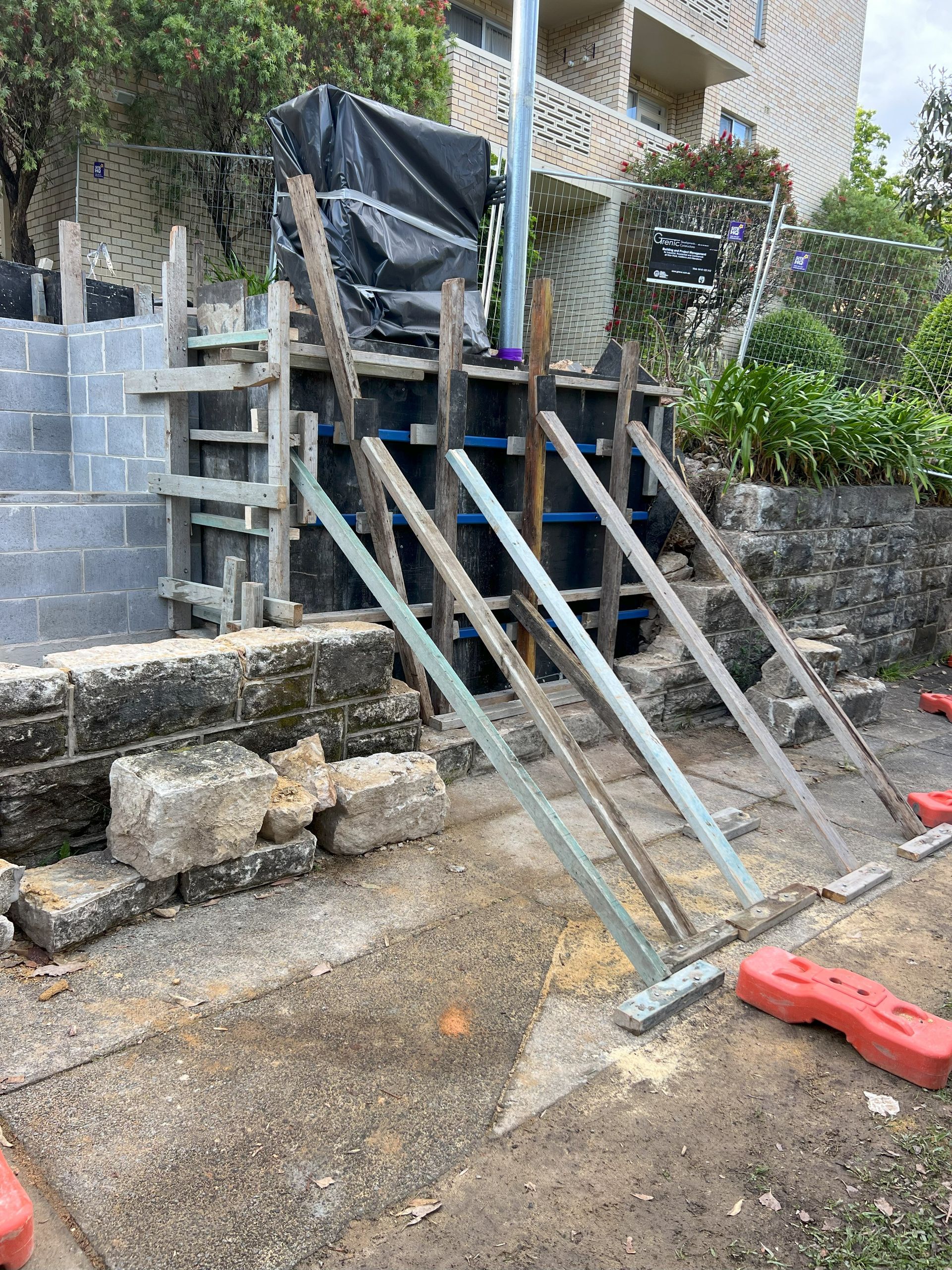 Construction site with concrete formwork, braced with timber and metal supports, on a sloped area next to a building.