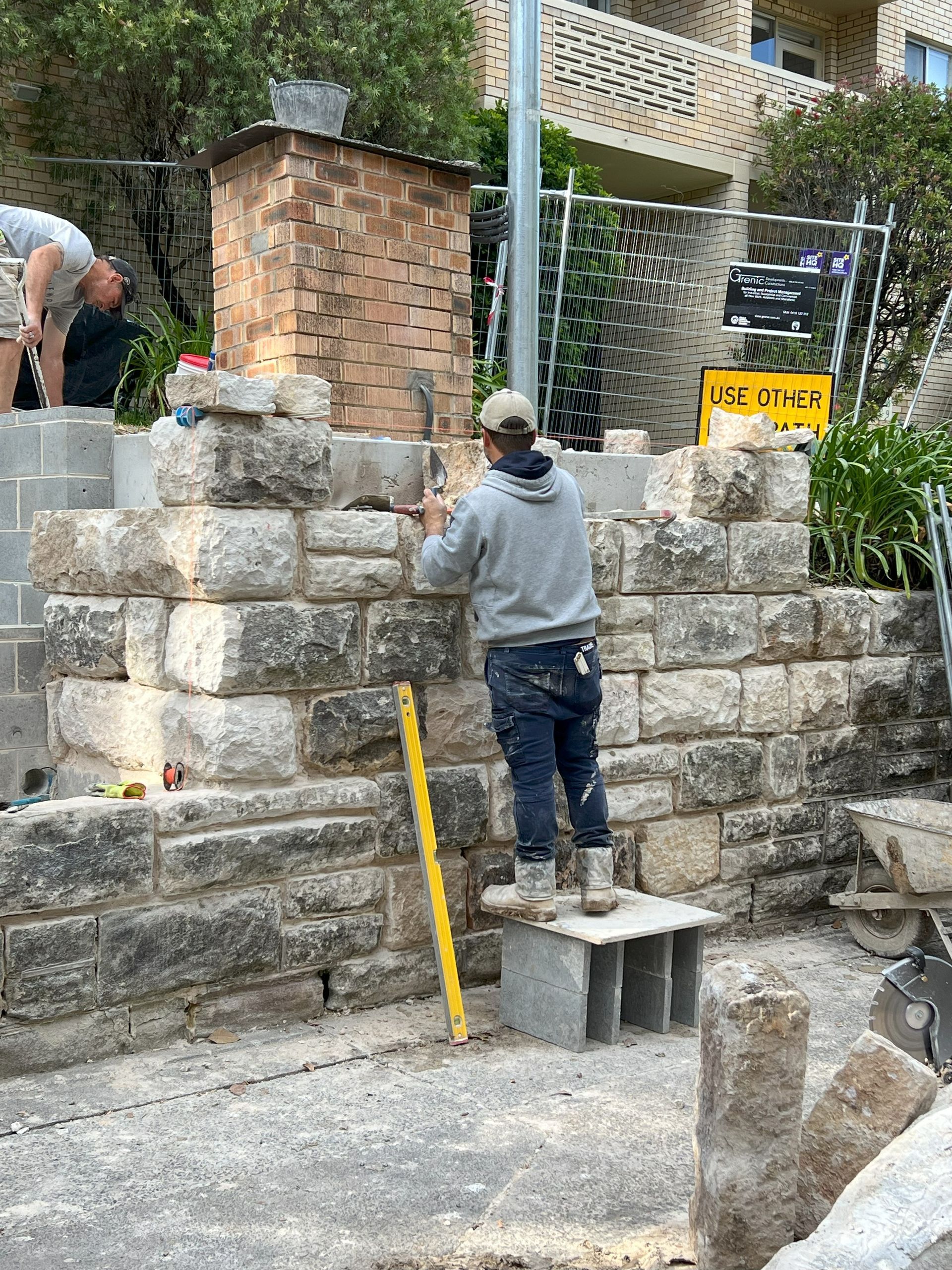 Construction worker building a stone wall with level and blocks; outdoor setting.