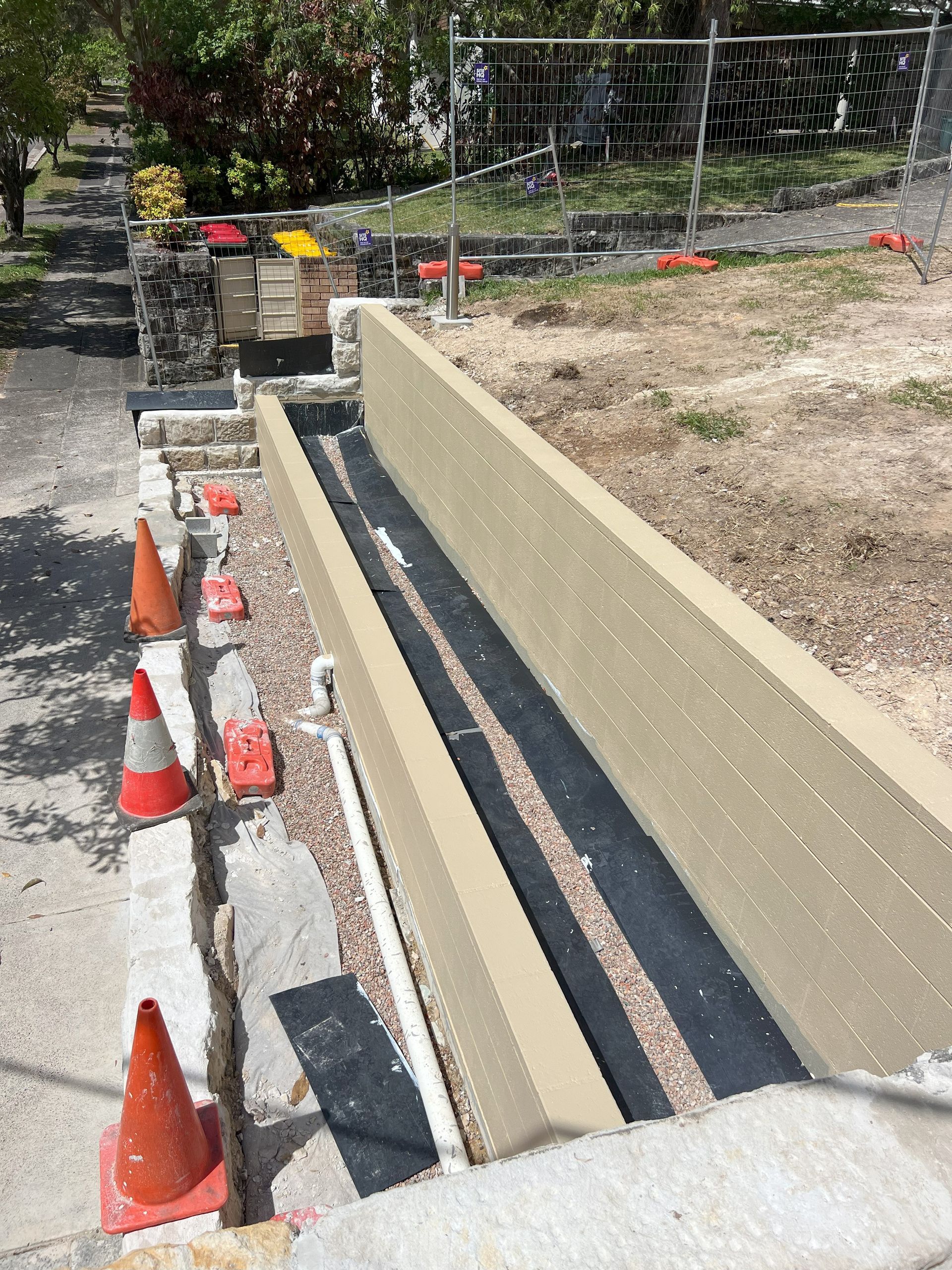 Construction site with a concrete drainage channel being built alongside a sidewalk. Orange cones and fencing are present.