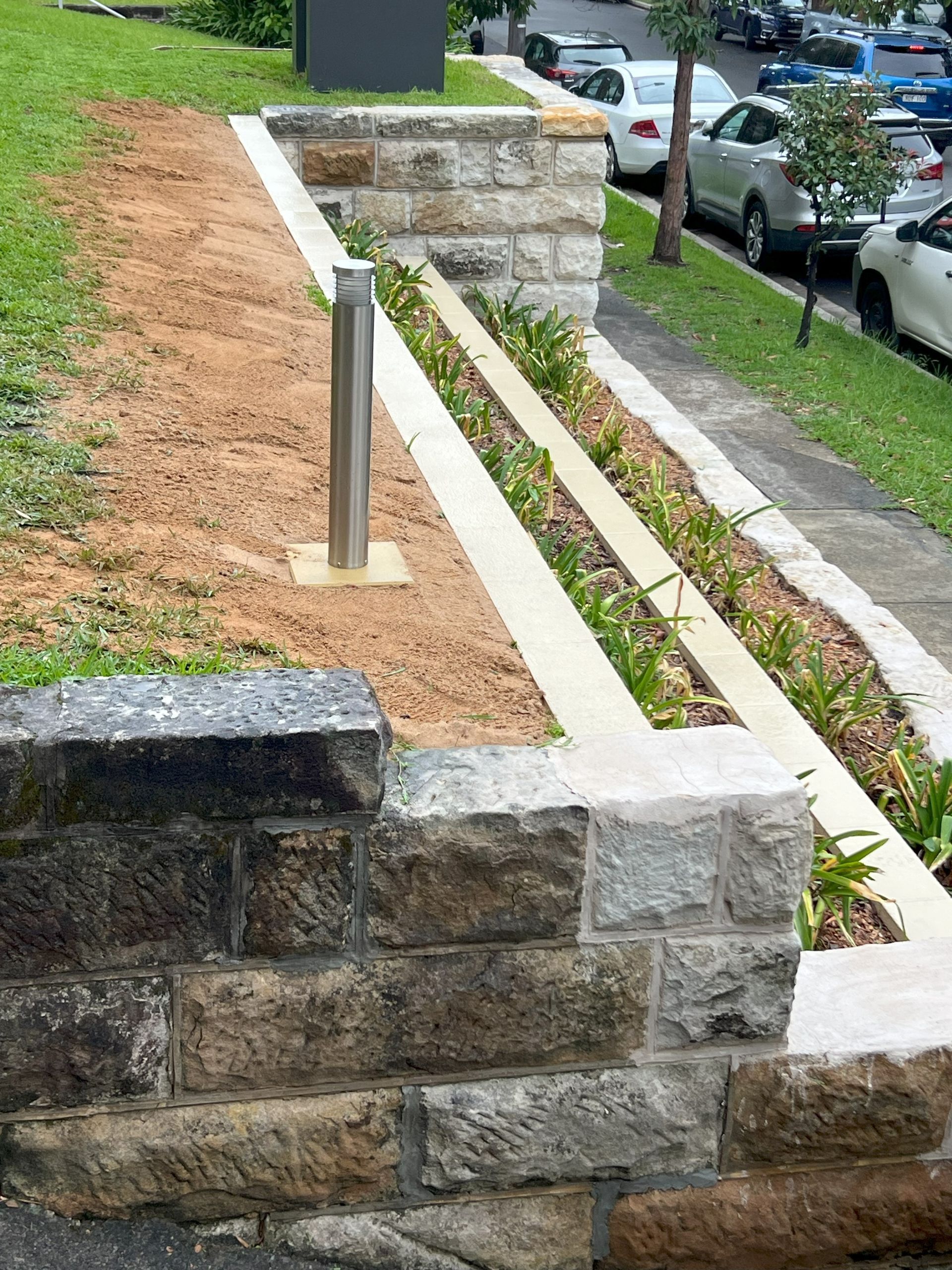 Stone retaining wall with a planting bed, and a bollard along a sidewalk, next to a street with parked cars.