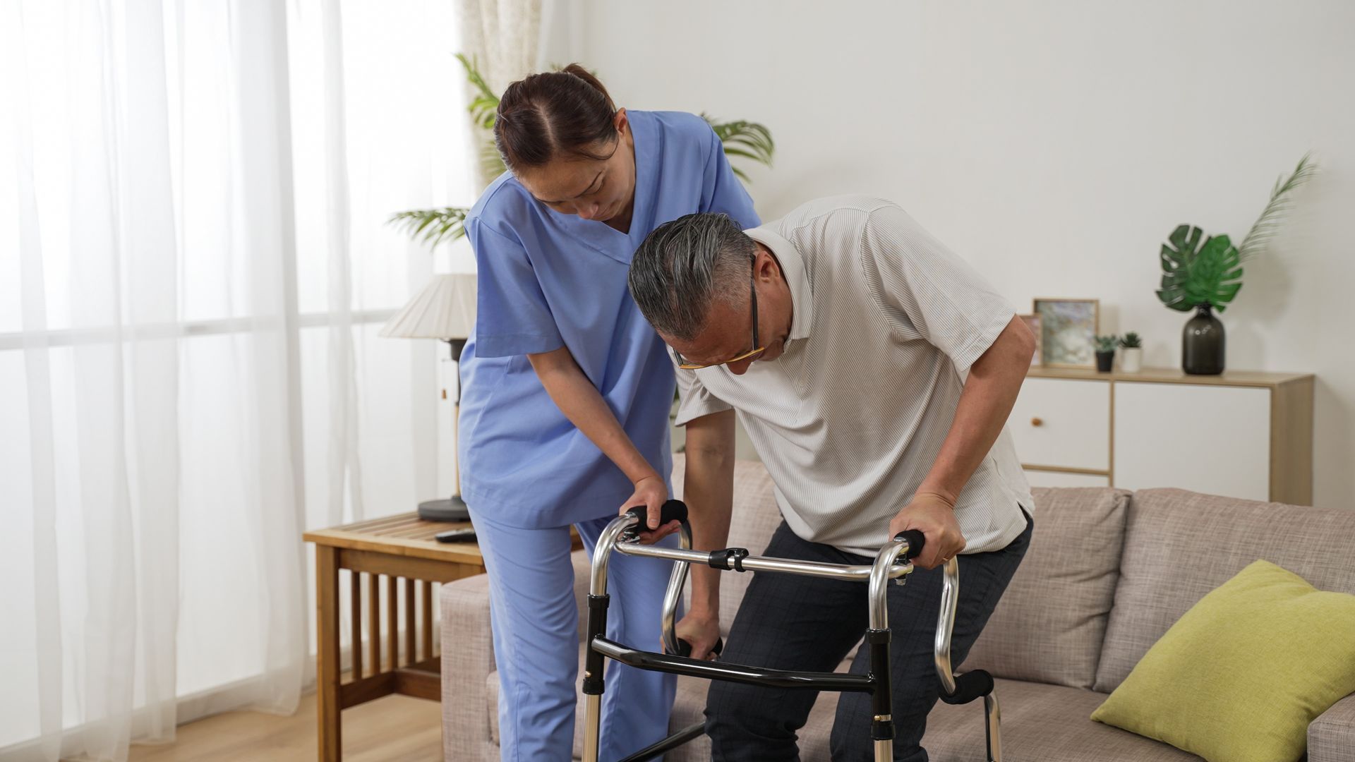 Caregiver assists an older adult using a walker in a living room.