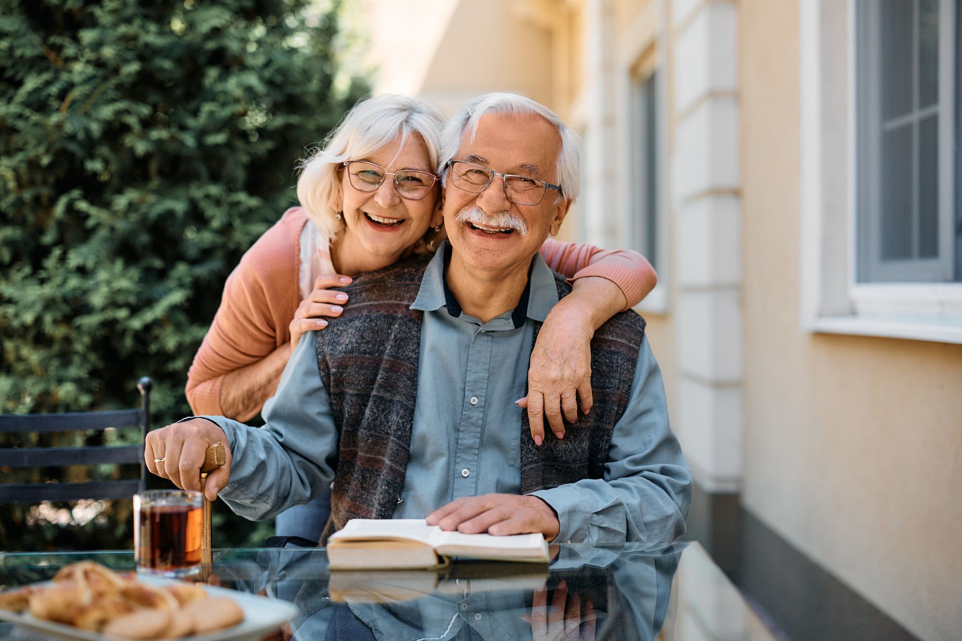 Smiling couple outdoors; woman's arms around man reading, both wearing glasses. Table with drink and food.