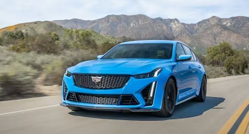 Blue Cadillac sedan driving on a road, mountains in the background.