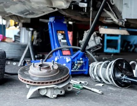 Car repair shop scene with a jack, suspension parts, and brake rotor on the floor.