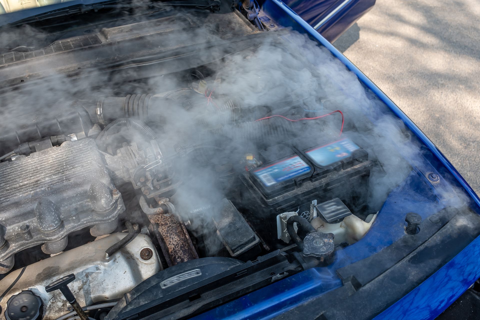 Thick white smoke billows from the open hood of a blue car, indicating an engine overheating or malfunction.
