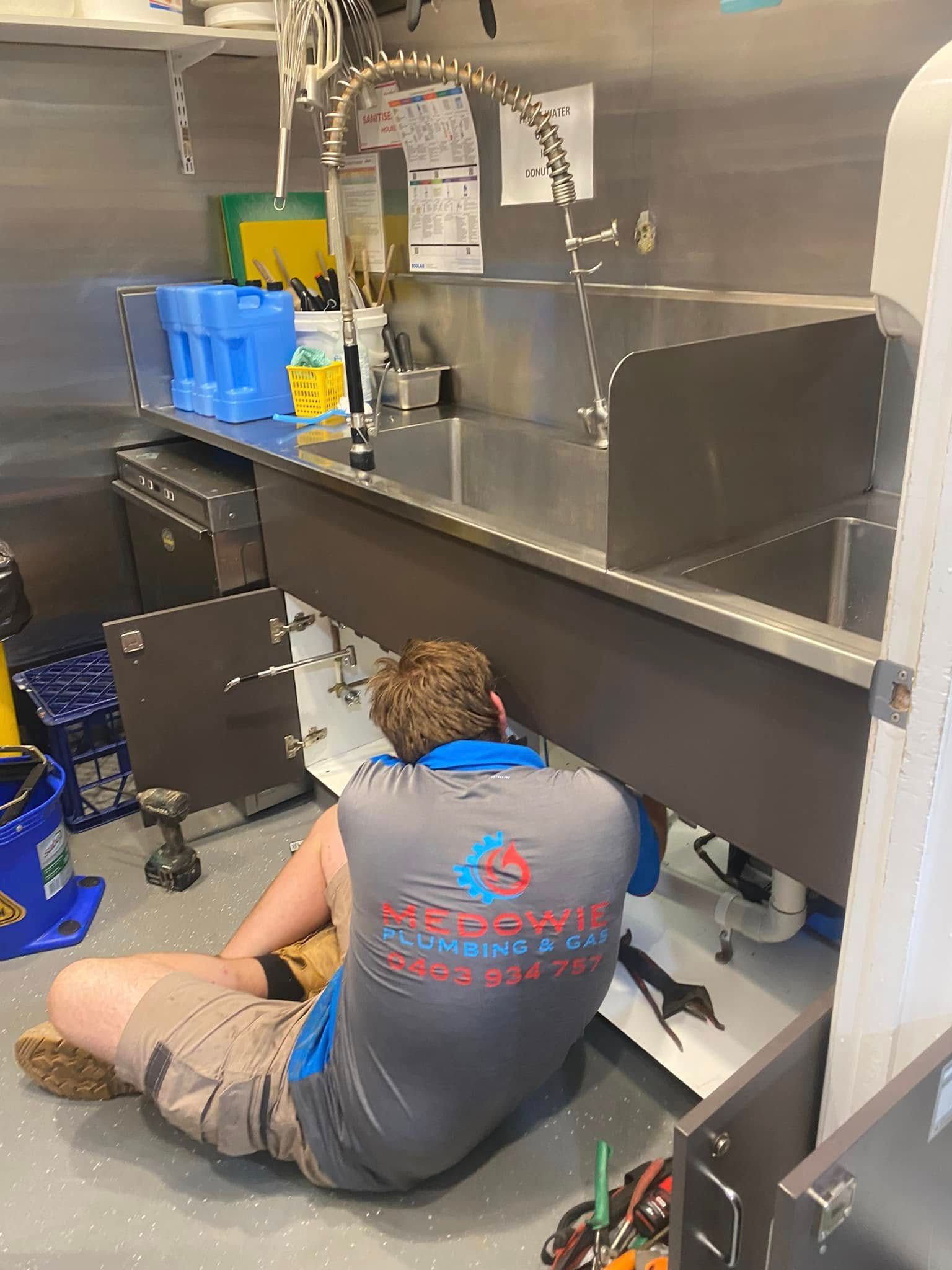 Person Sitting on the Floor in a Commercial Kitchen Under a Stainless-steel Counter — Medowie Plumbing & Gas in in Maitland