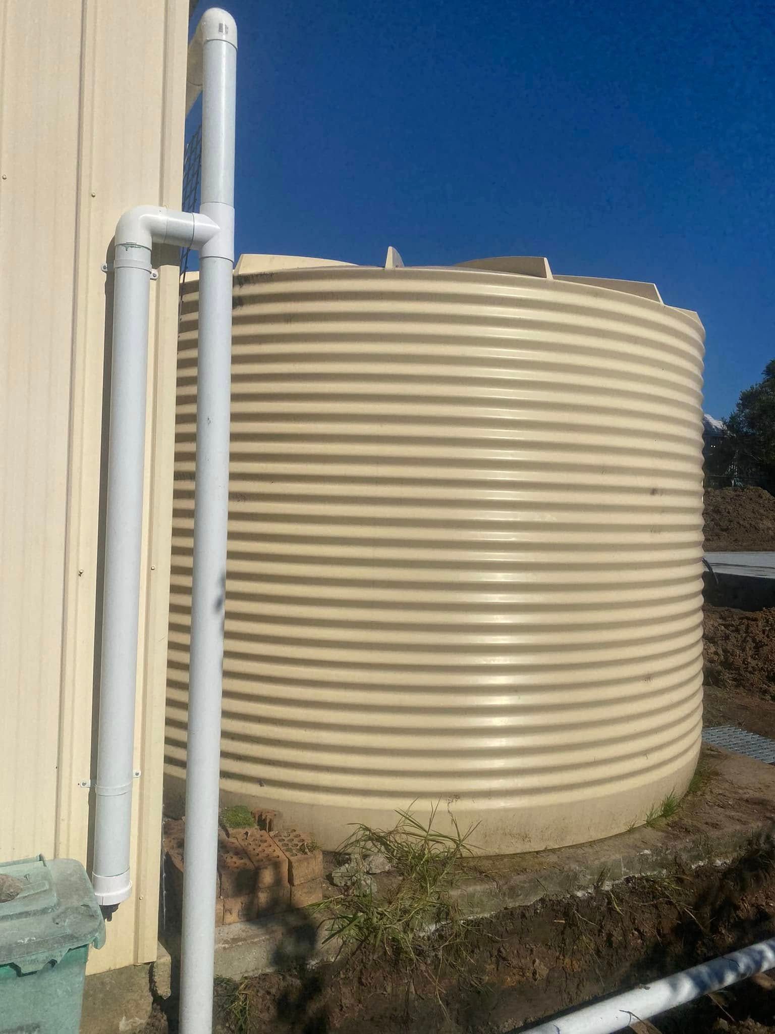 Large beige ribbed water tank beside a building with white pipes under a clear blue sky — Medowie Plumbing & Gas In Medowie, NSW