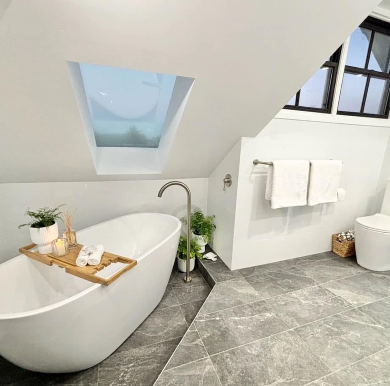 Modern white attic bathroom with a freestanding tub, skylight, grey tile floor, and towel rack — Medowie Plumbing & Gas In Medowie, NSW