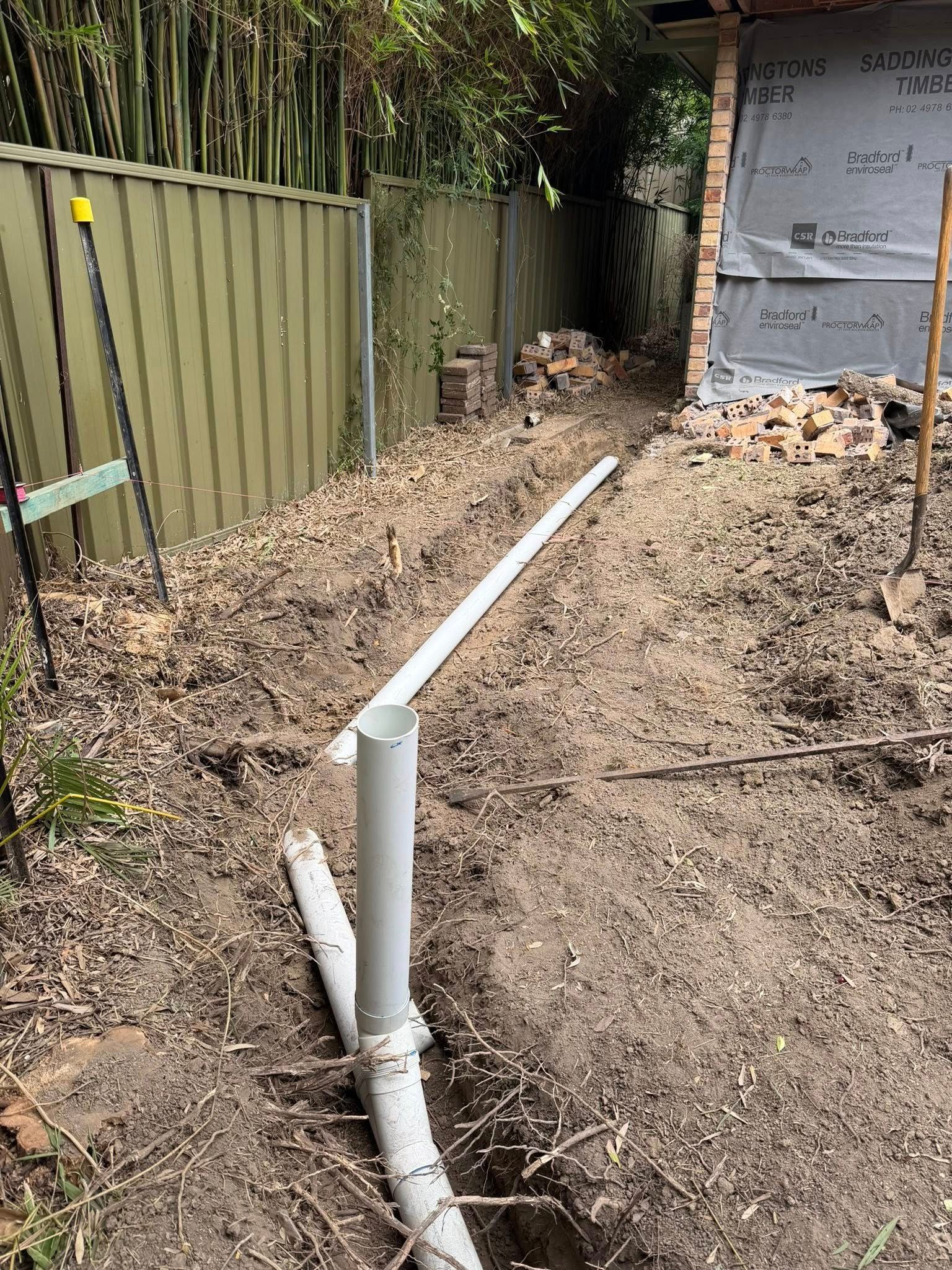Narrow dirt path beside a green fence, with white pipes and debris leading toward a work area— Medowie Plumbing & Gas In Medowie, NSW