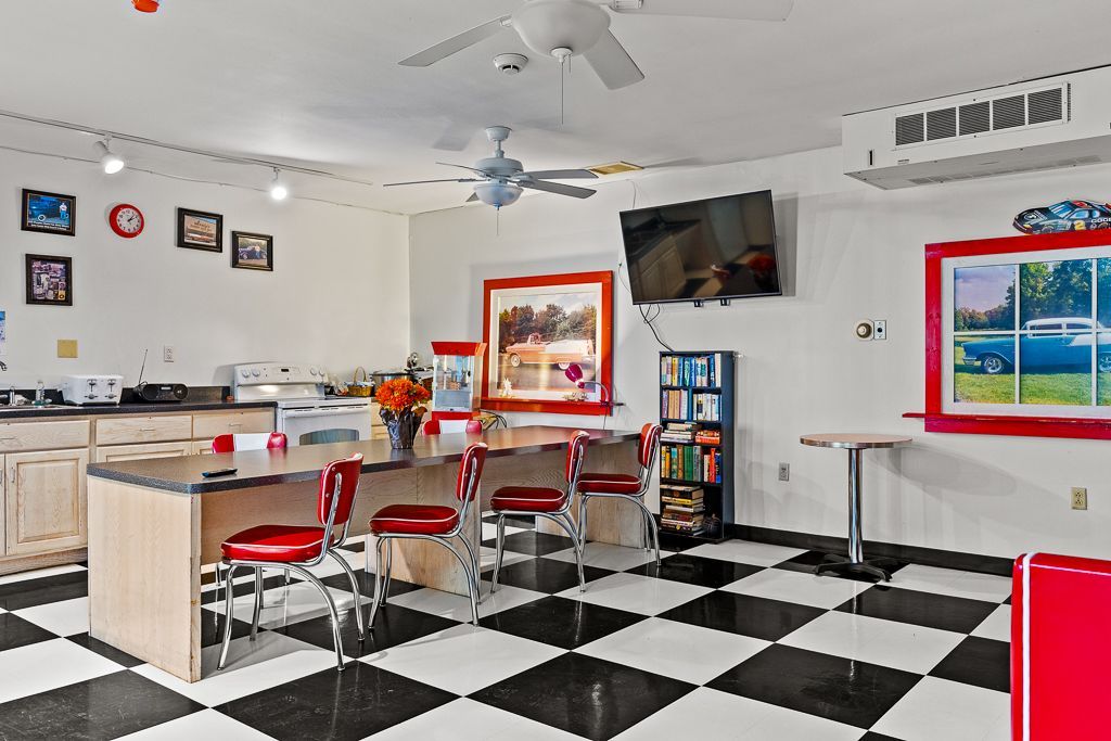 Retro diner-themed room with a black and white checkered floor, kitchen, seating area, and framed art.