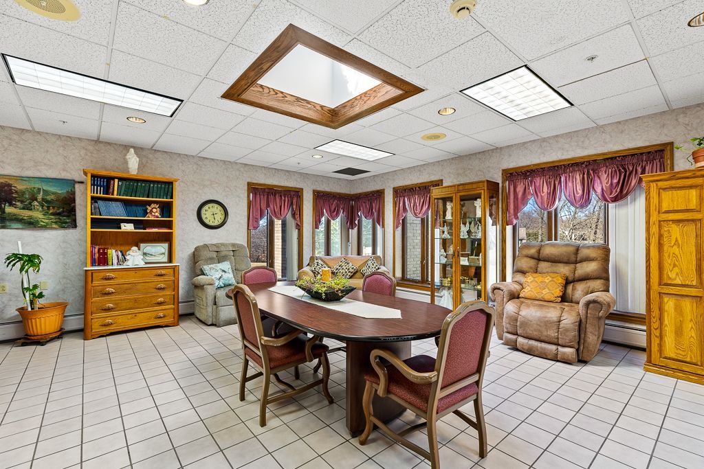 Brightly lit dining/living room in a senior care facility with seating, a large table, and cabinetry.