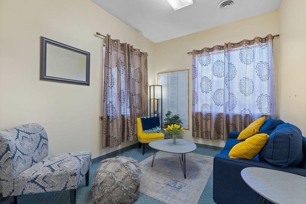 Living room with blue sofa, yellow chair, patterned armchair, mirror, curtains, rug, and small tables.